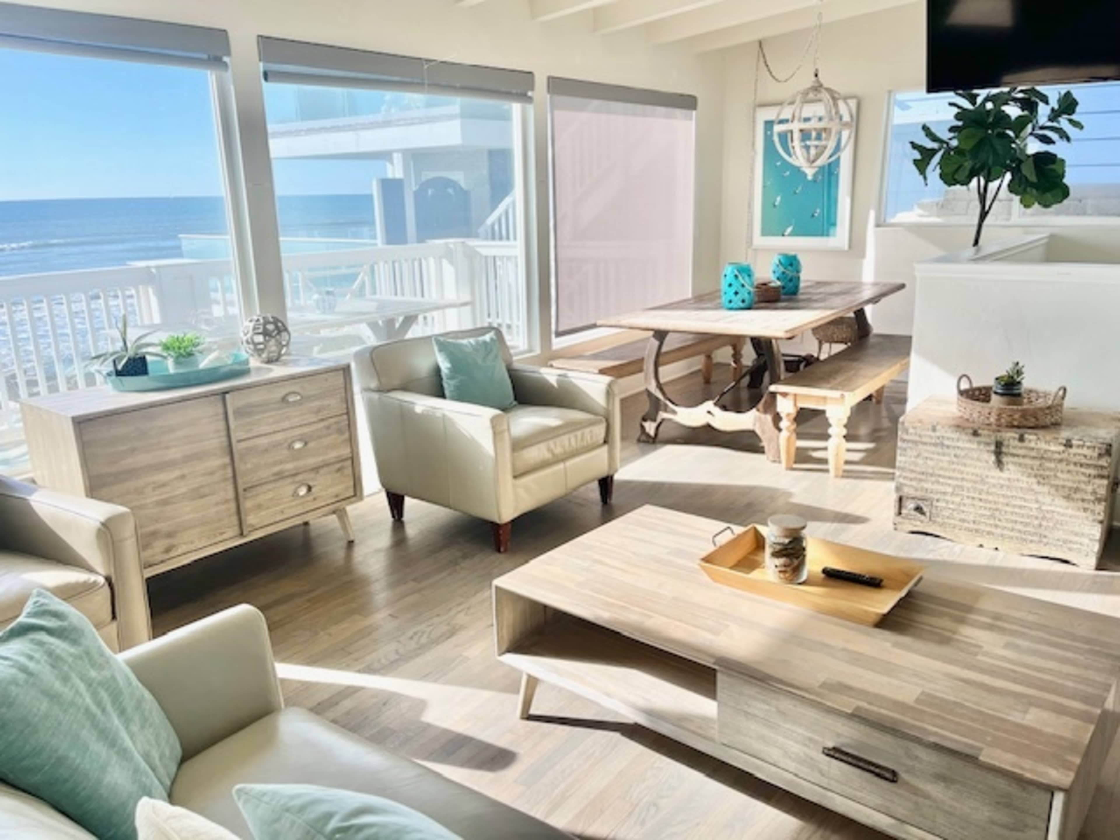 A coastal living room features large windows with a view of the ocean, a wooden dining table, and light-colored furniture.