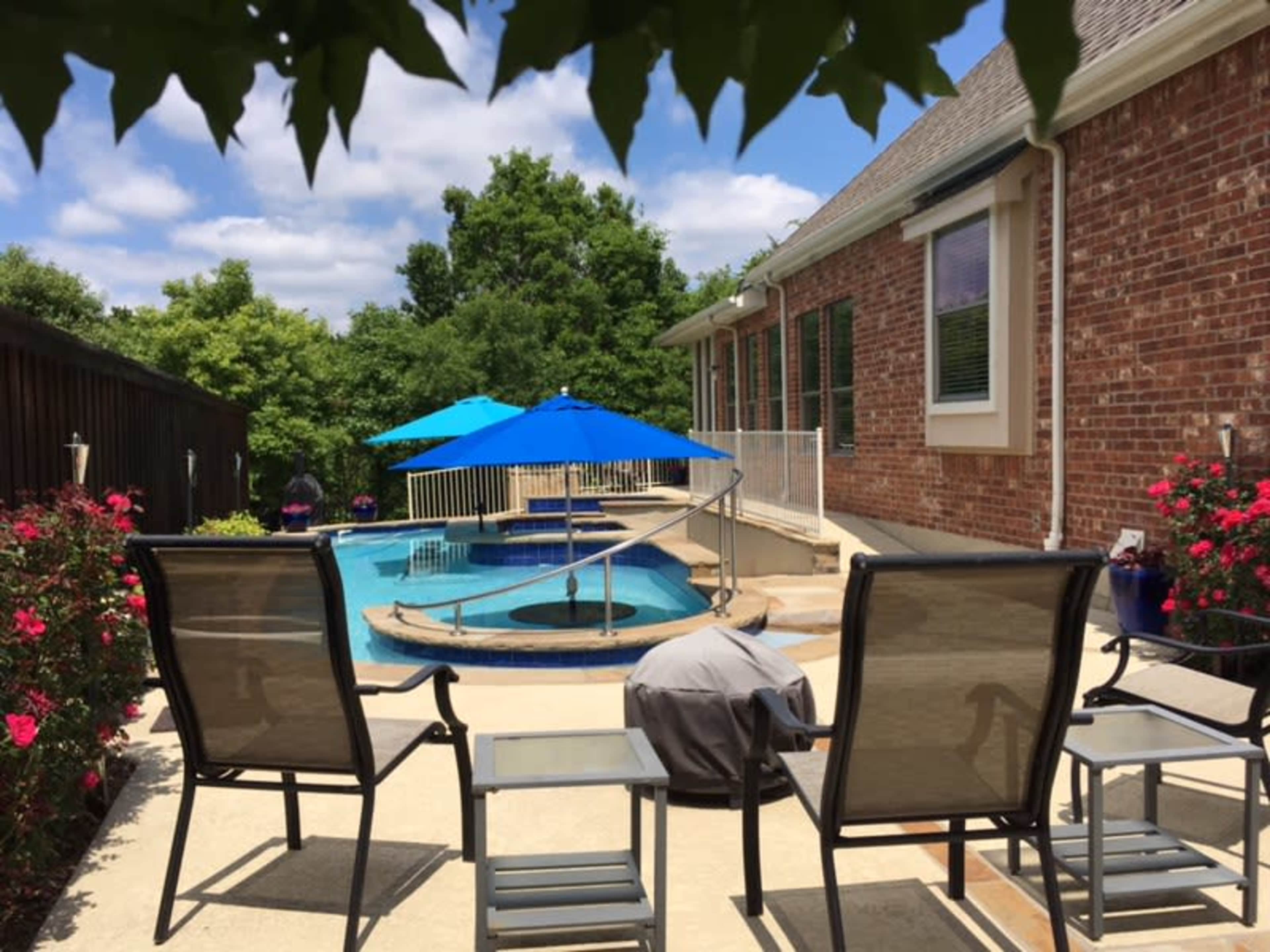 A patio features several chairs facing a blue pool surrounded by greenery and a brick house.