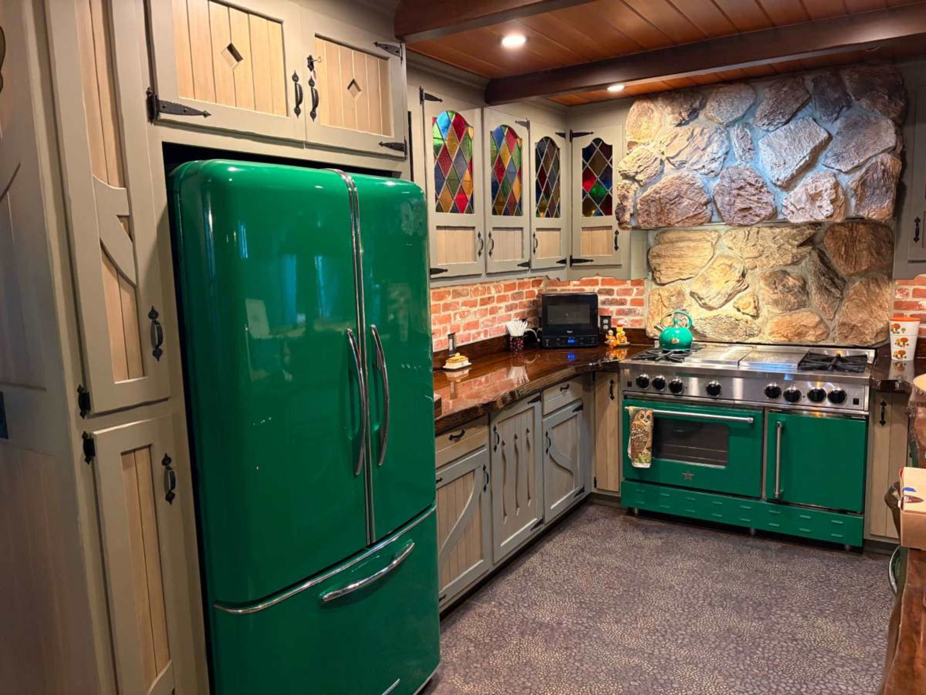 The kitchen features green appliances, including a refrigerator and stove, set against a backdrop of stone walls and wooden cabinetry.