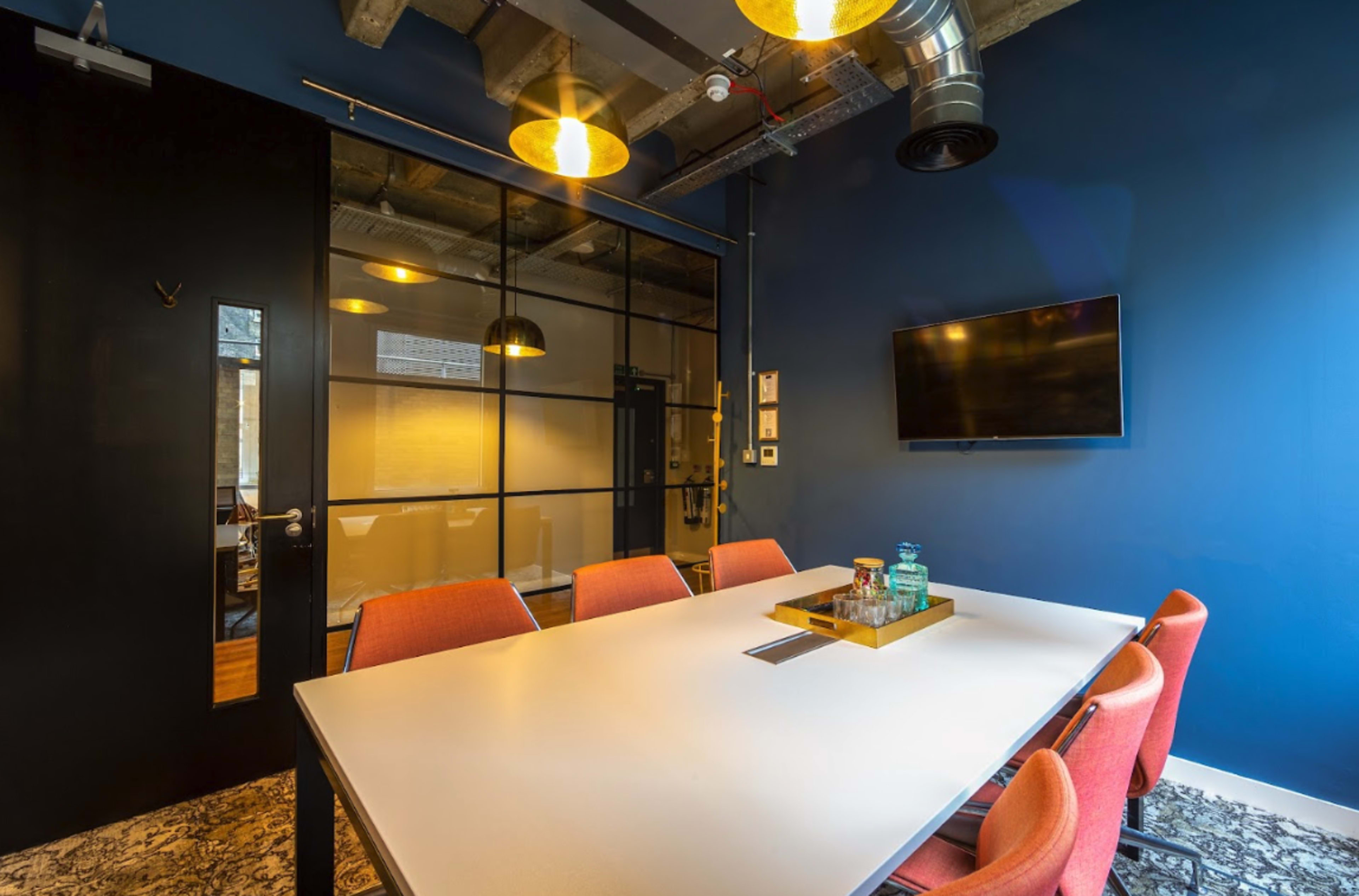 The image shows a modern conference room with a white table, orange chairs, a glass wall, and a wall-mounted television.