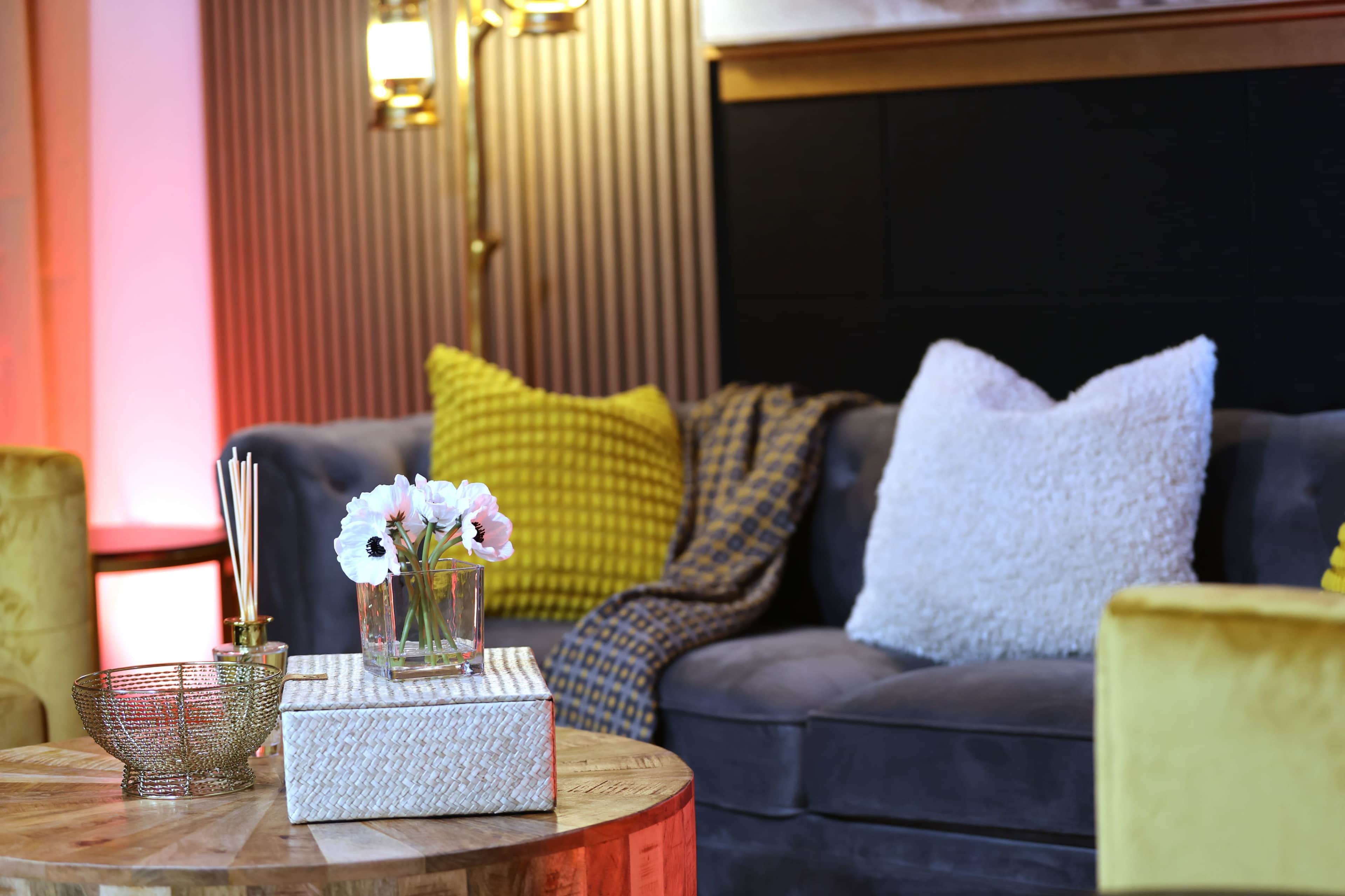 A cozy living room scene features a gray sofa adorned with yellow and white pillows, a wooden coffee table with decorative items, and a small vase of flowers.