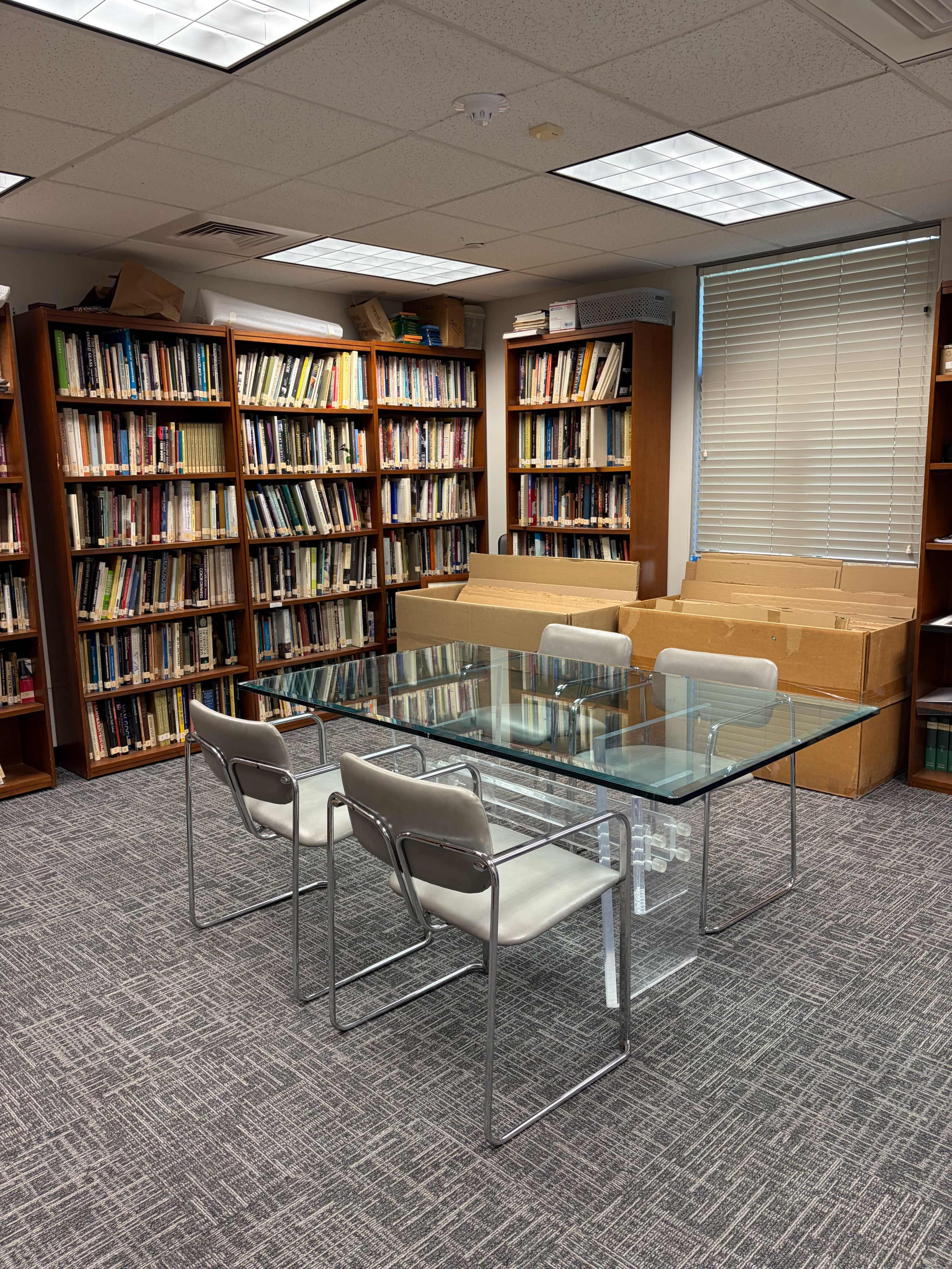 The image shows a library room with a glass table and four chairs surrounded by shelves filled with books, while cardboard boxes are stacked in one corner.