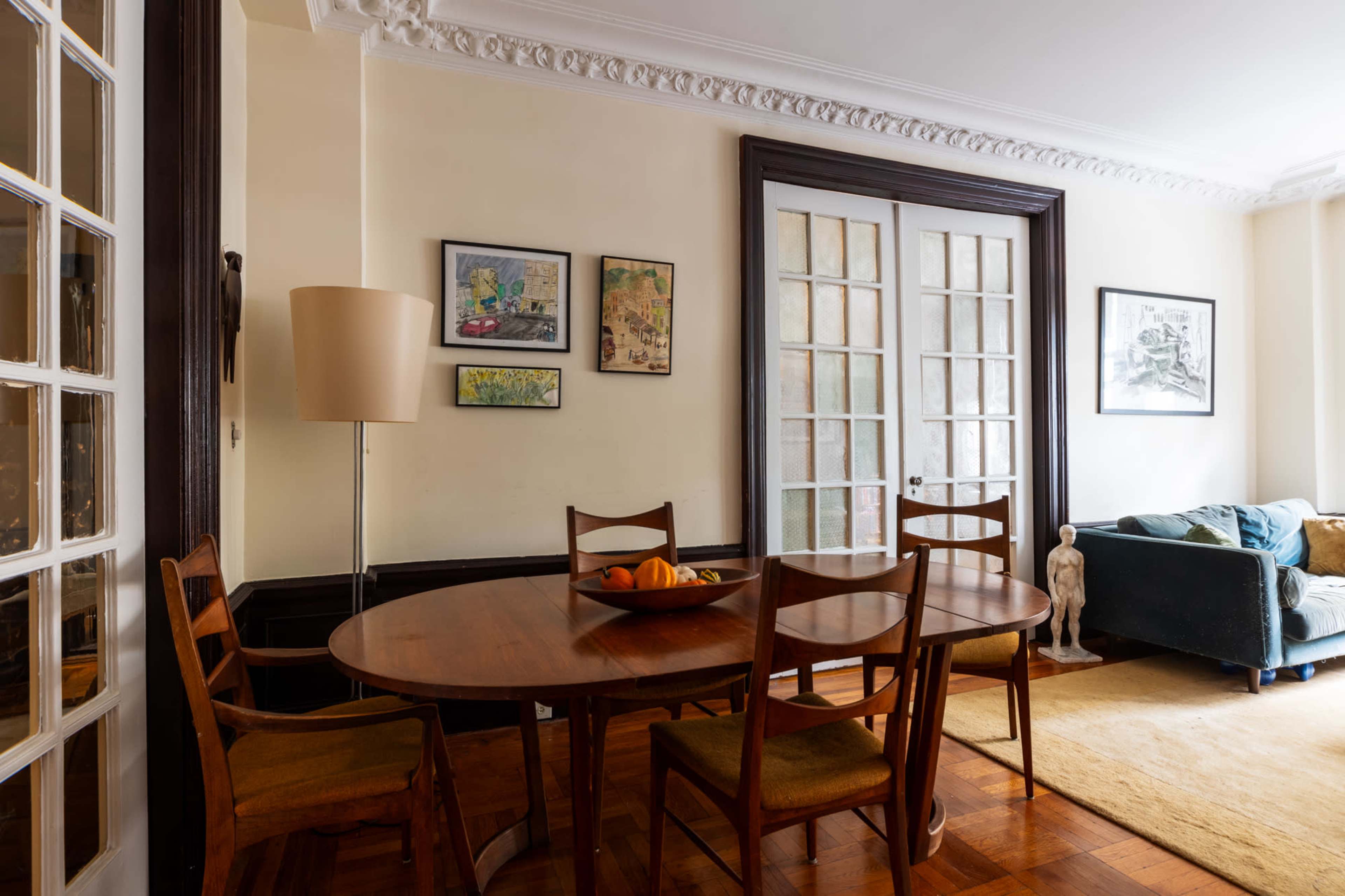The image shows a dining area with a wooden table and four chairs, framed artwork on the walls, a floor lamp, and a living room visible through glass doors.