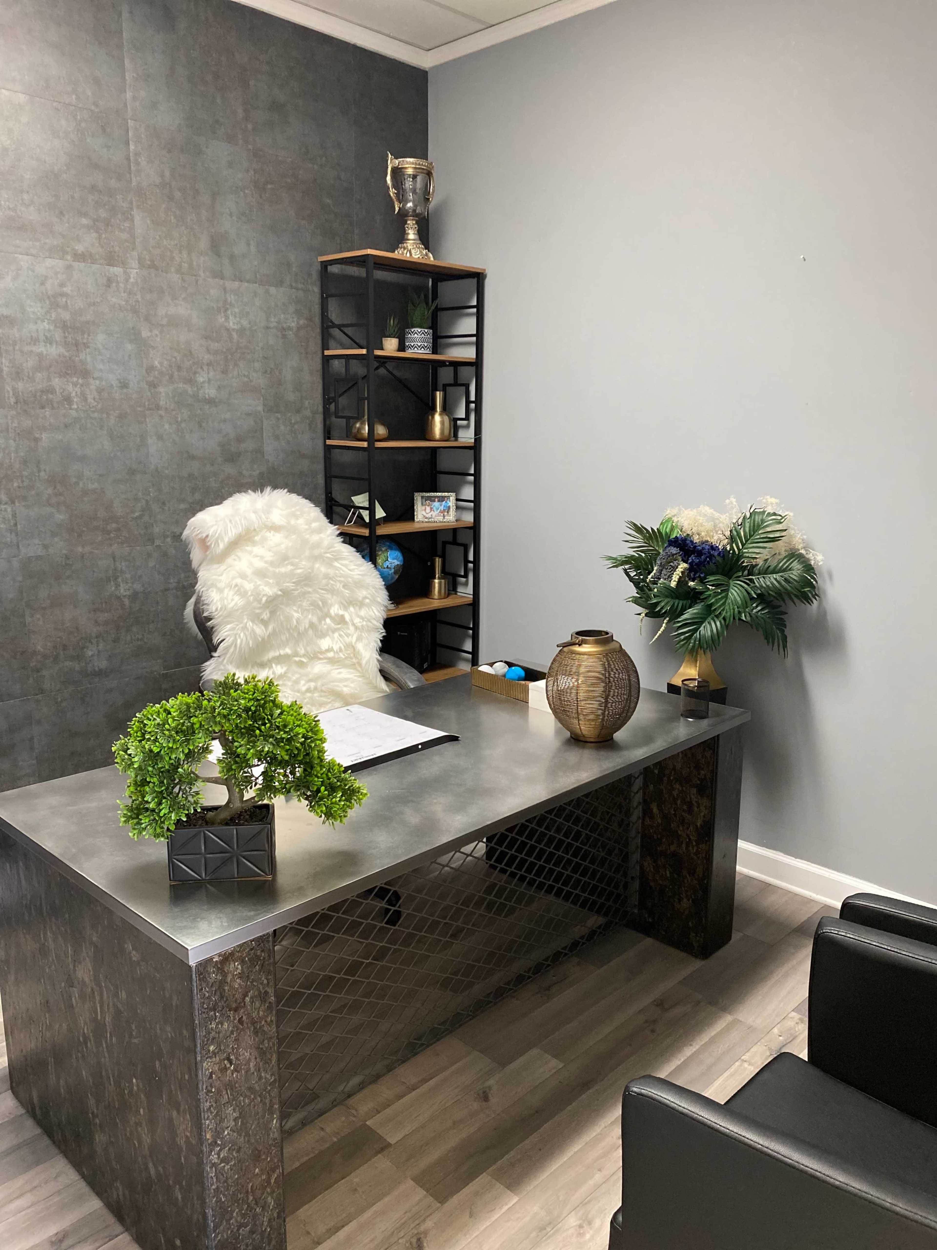 The image shows a modern office space with a metal desk, a plush white chair, a bookshelf with decorative items, and a small arrangement of plants and flowers.