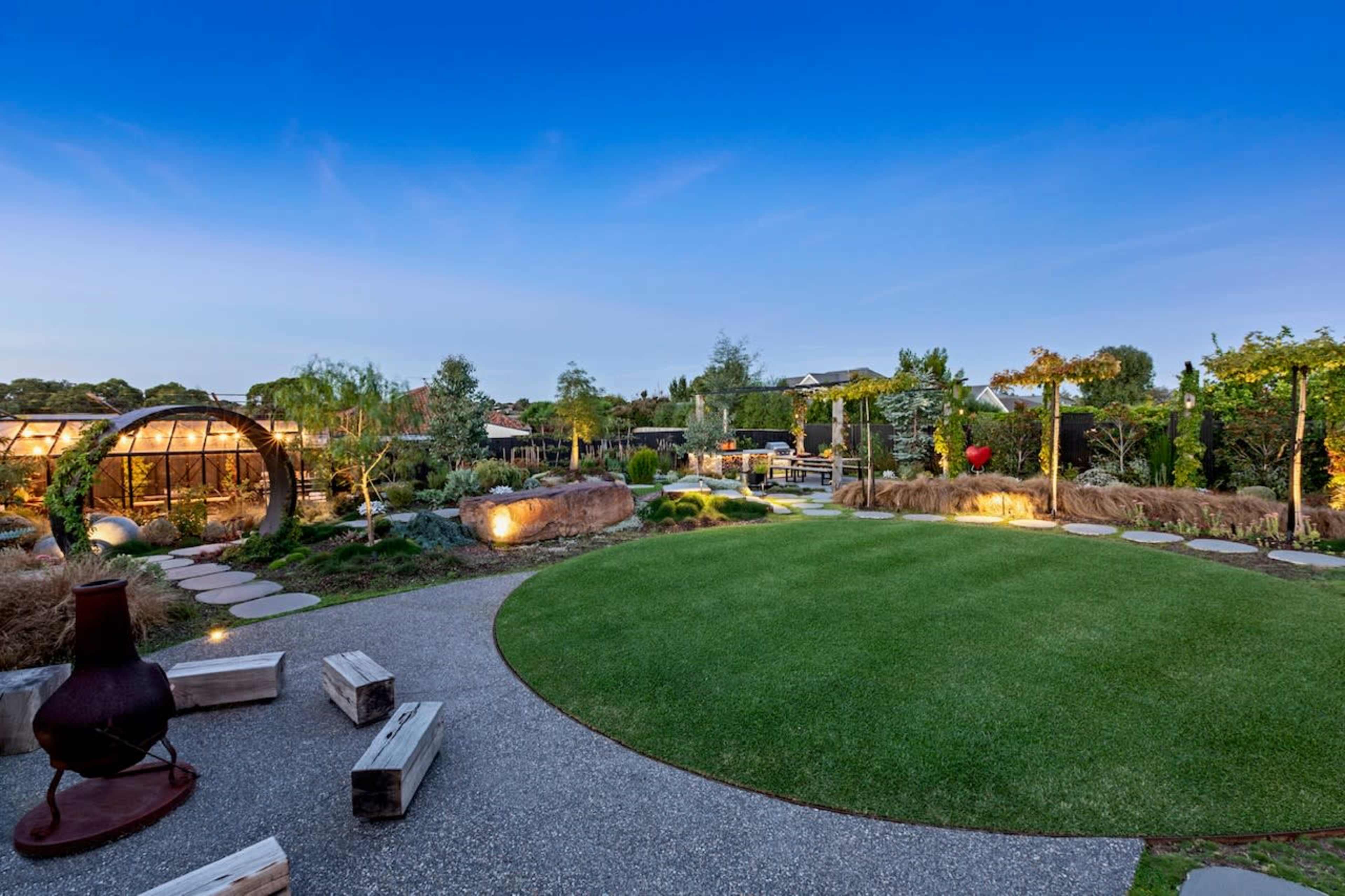 A landscaped garden with a curved pathway, seating areas made of logs, and a textured lawn under a clear blue sky.