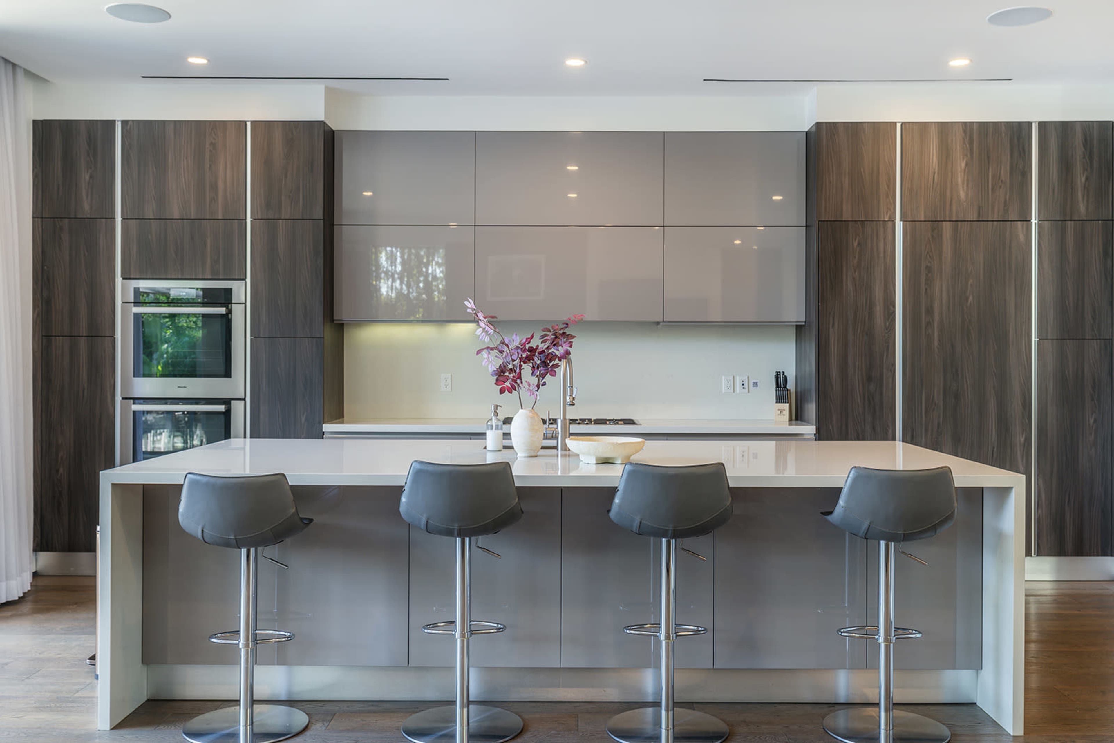 The image features a modern kitchen with a central island and four high-backed stools, complemented by sleek cabinetry and appliances.