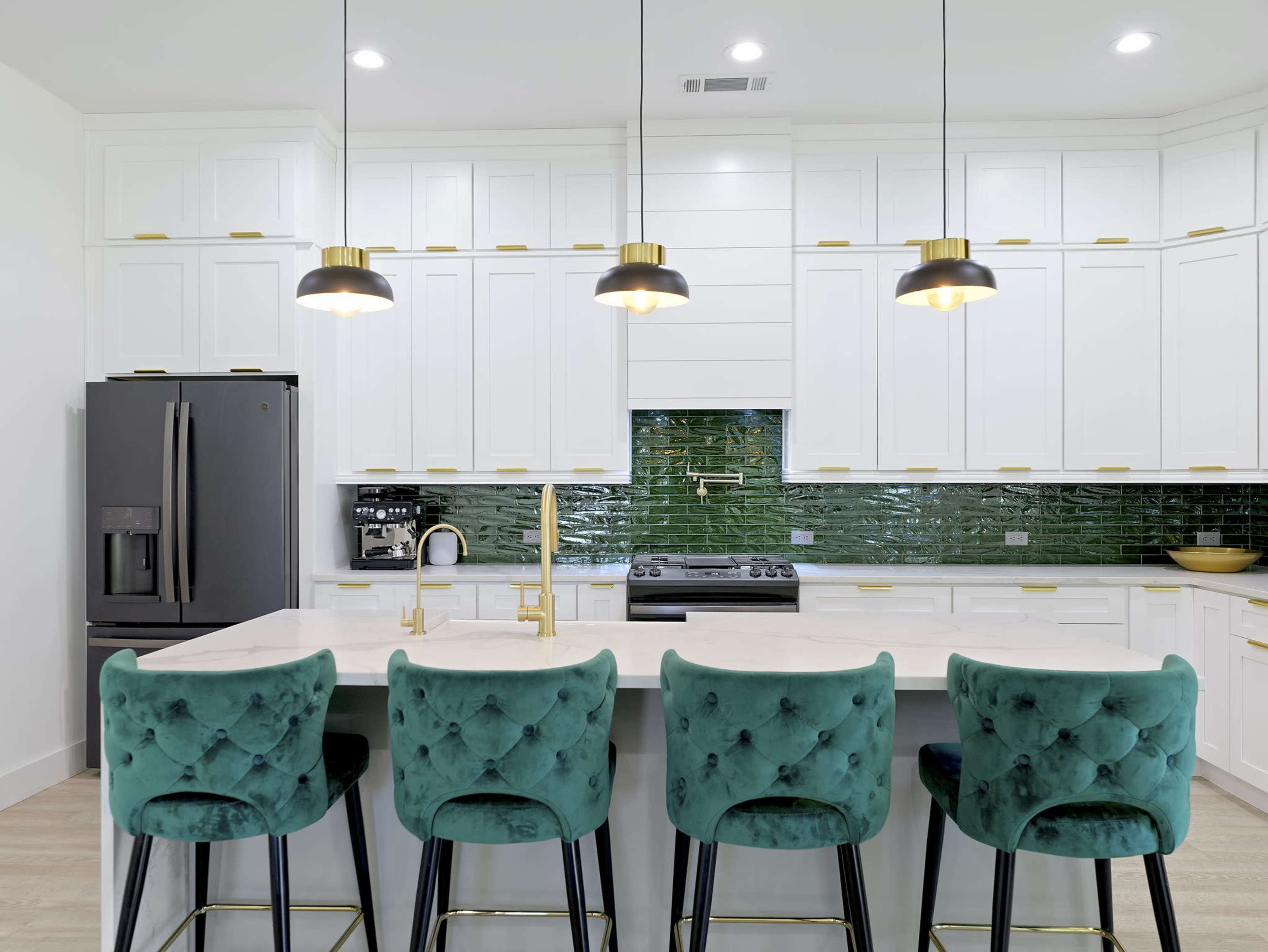 The image shows a modern kitchen featuring white cabinetry, green tiled backsplash, and four teal upholstered stools at a white countertop.