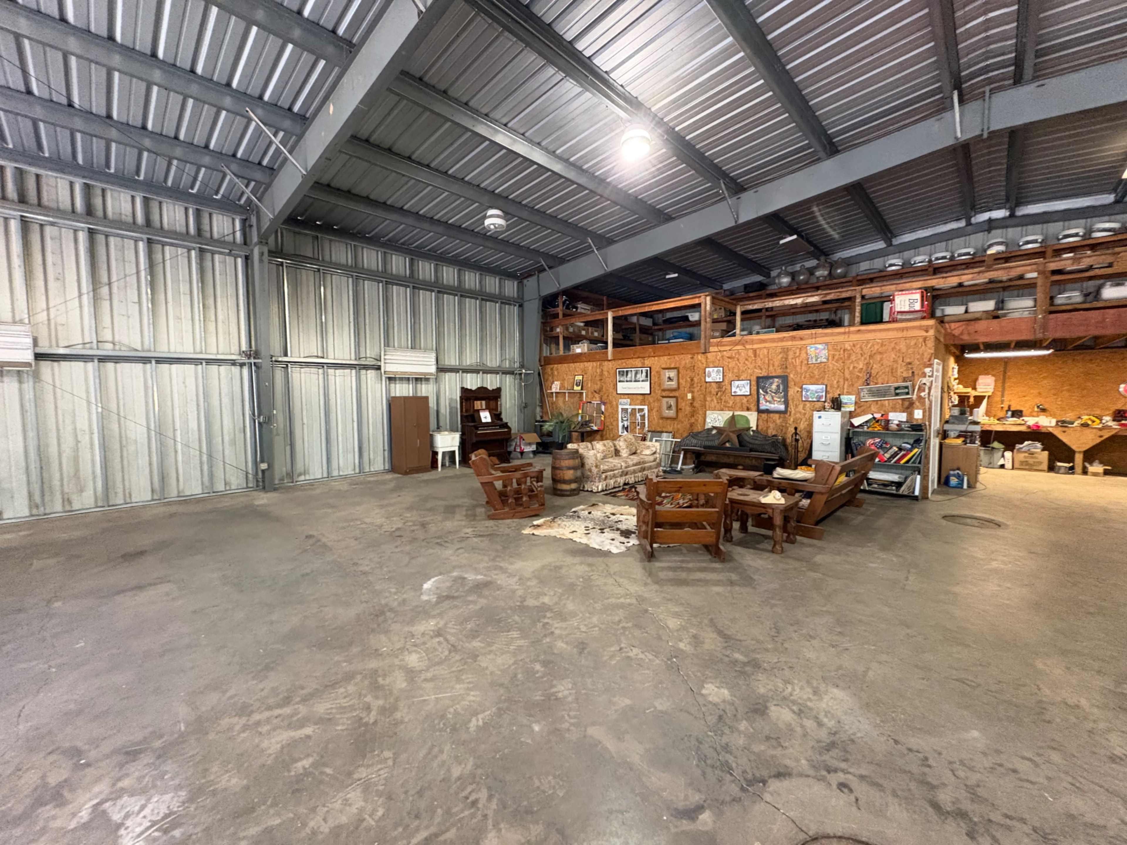 The image shows a spacious, industrial-style interior with a concrete floor, metal framework, and wooden furniture arranged in a seating area.