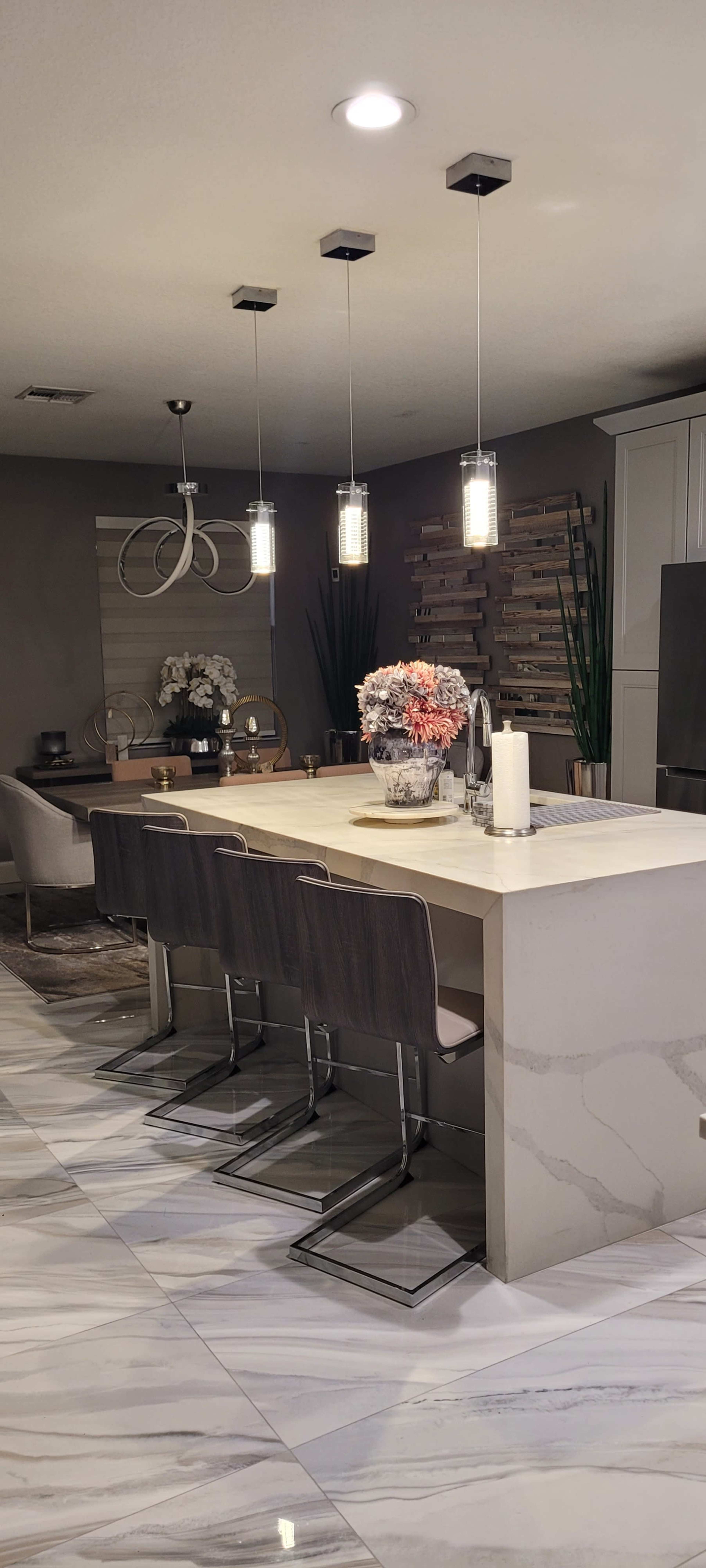 A modern kitchen features a large island with seating, pendant lights overhead, and a decorative arrangement of flowers and plants.