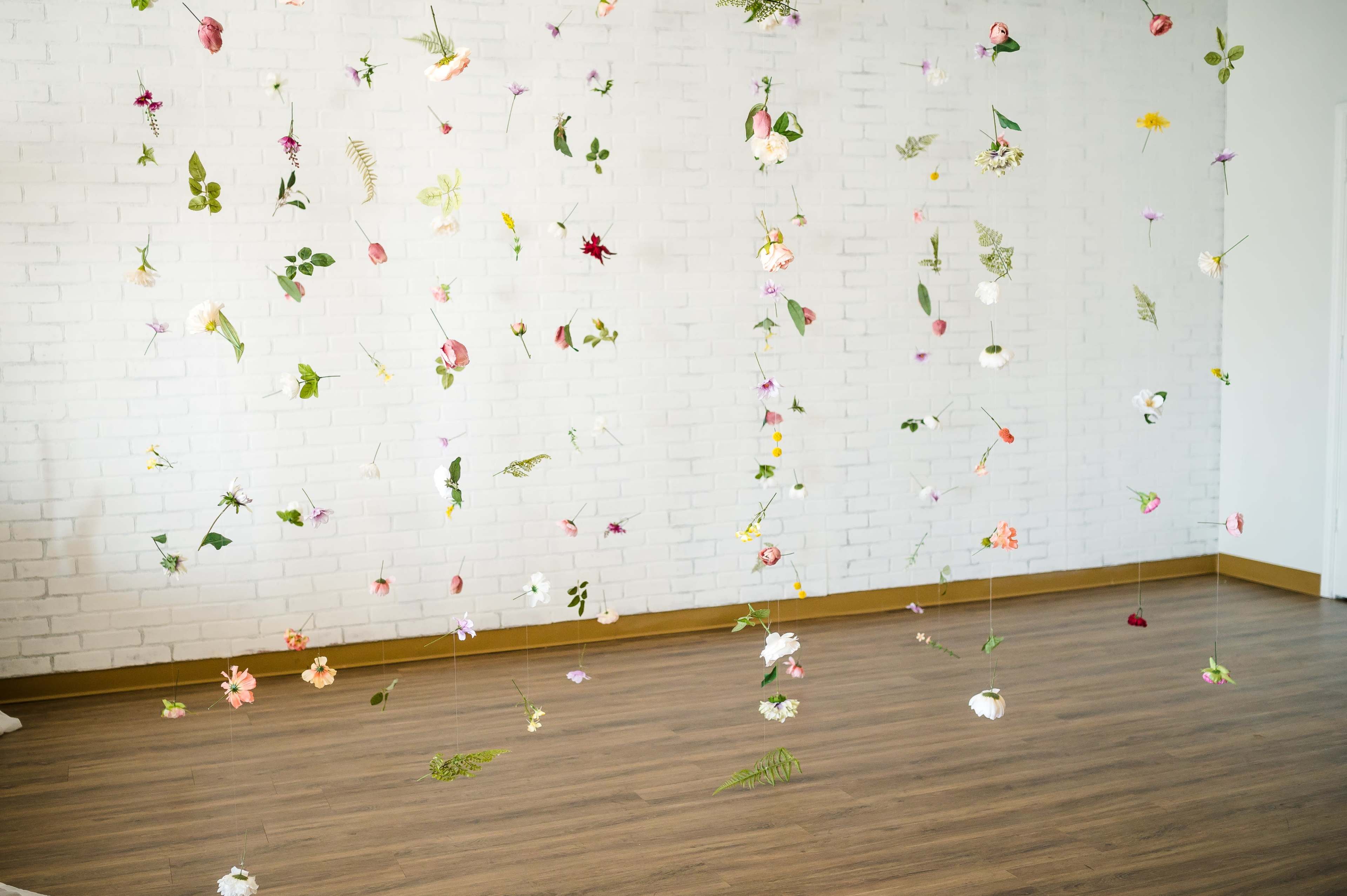 A collection of suspended flowers and leaves hangs from the ceiling in a bright, empty room with a white brick wall and wooden floor.