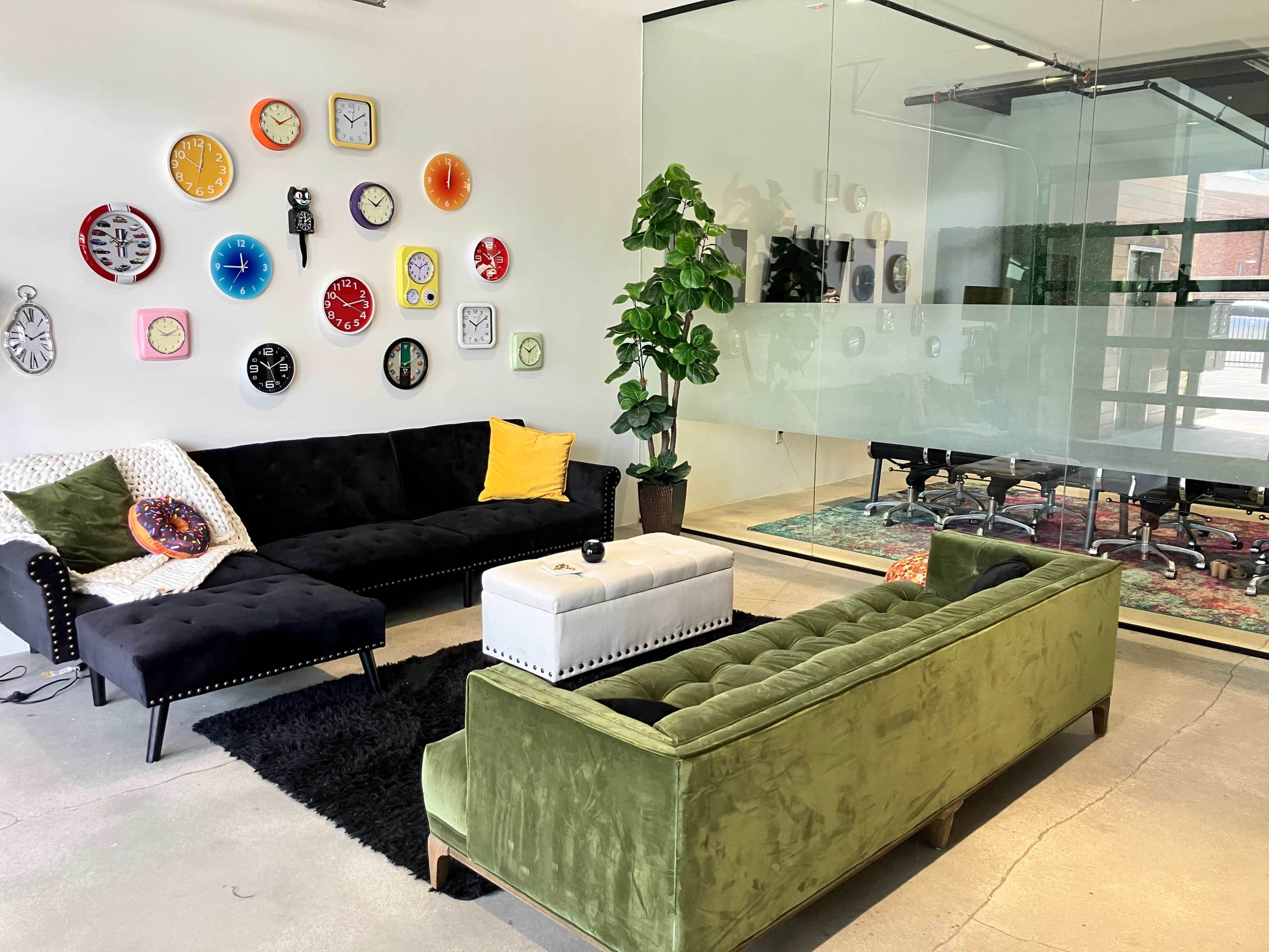 The scene shows a modern lounge area with two sofas, various wall clocks, a potted plant, and a glass-walled meeting room in the background.