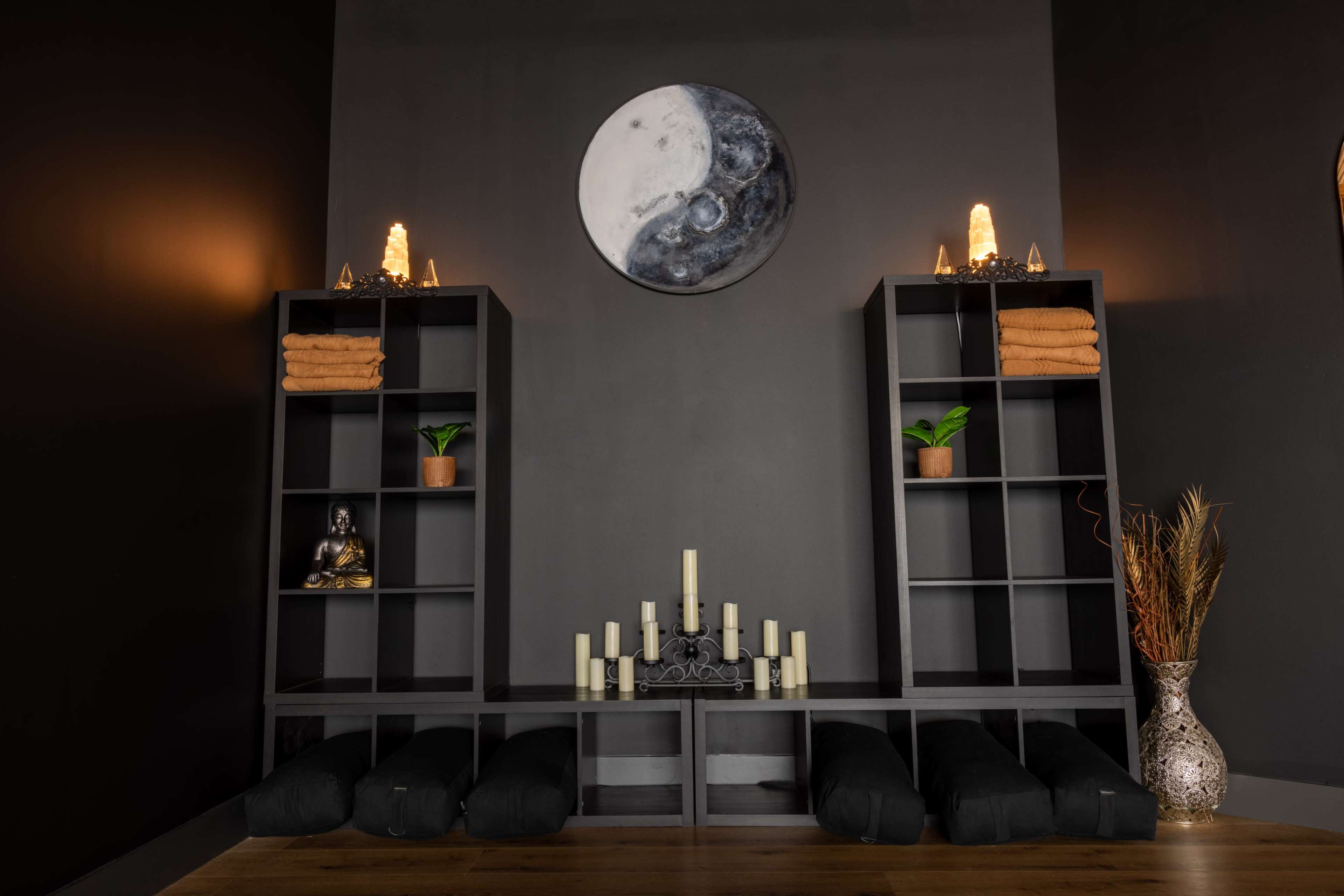 A serene room arranged with two dark shelving units, neatly stacked towels, a Buddha statue, candles, and plants, beneath a circular yin and yang wall art.