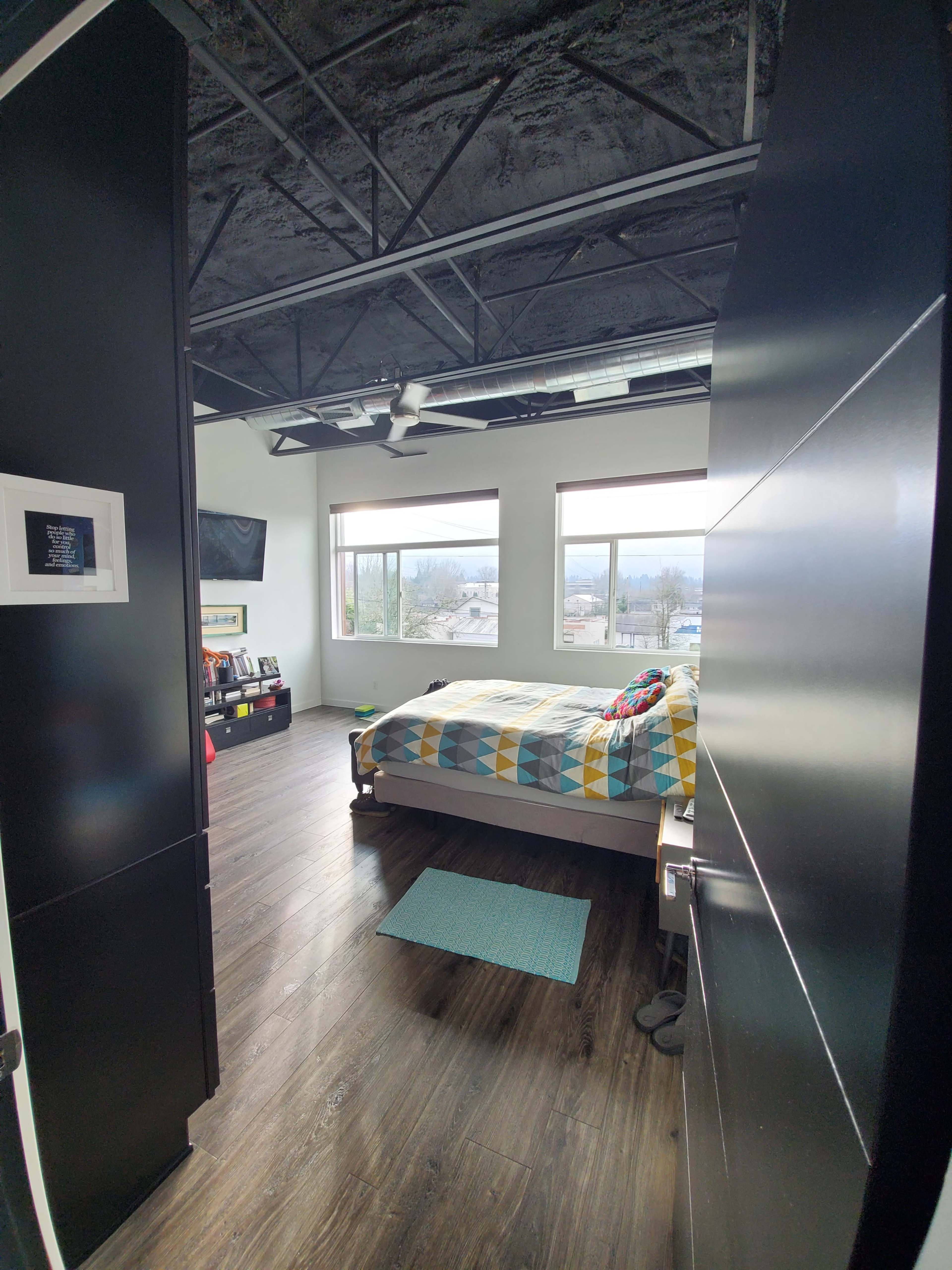 A bedroom is visible through an open doorway, featuring a bed with a patterned quilt, two windows, and a dark ceiling.