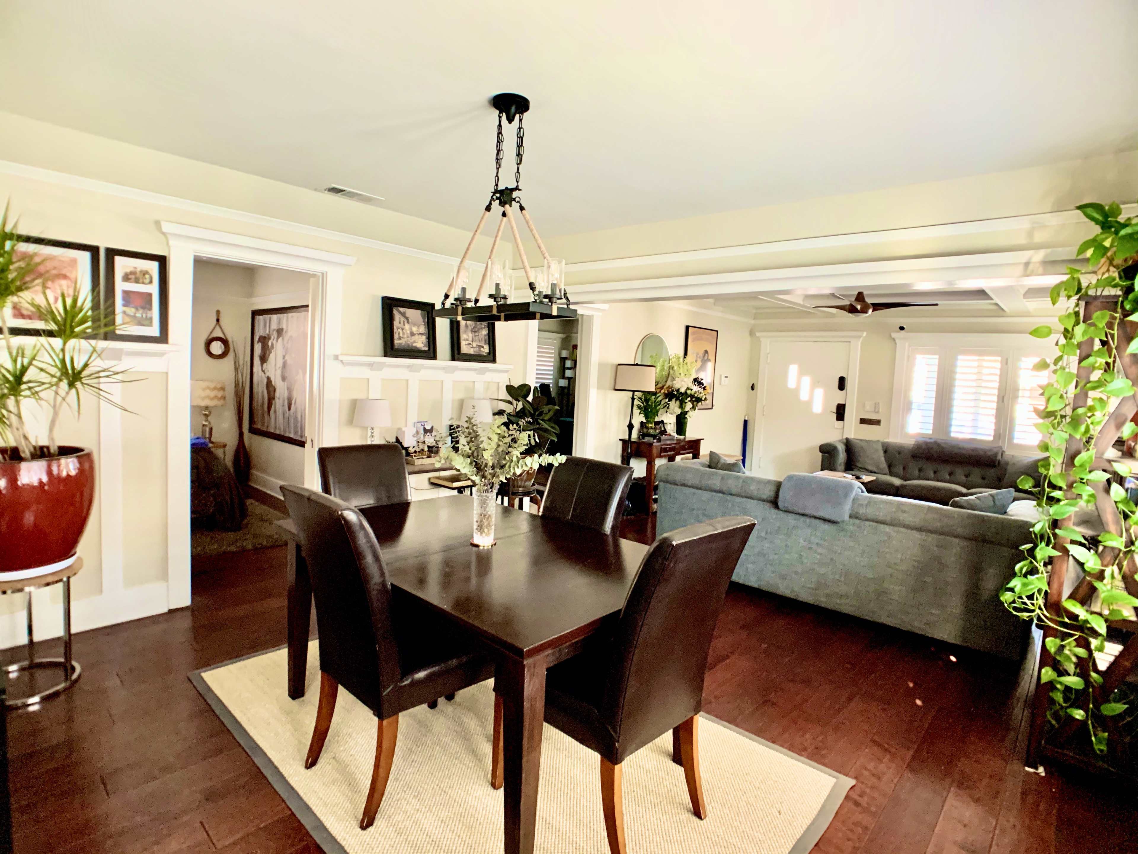 A dining area with a dark wooden table and black chairs, adjacent to a light-colored living room featuring a blue couch and decorative plants.