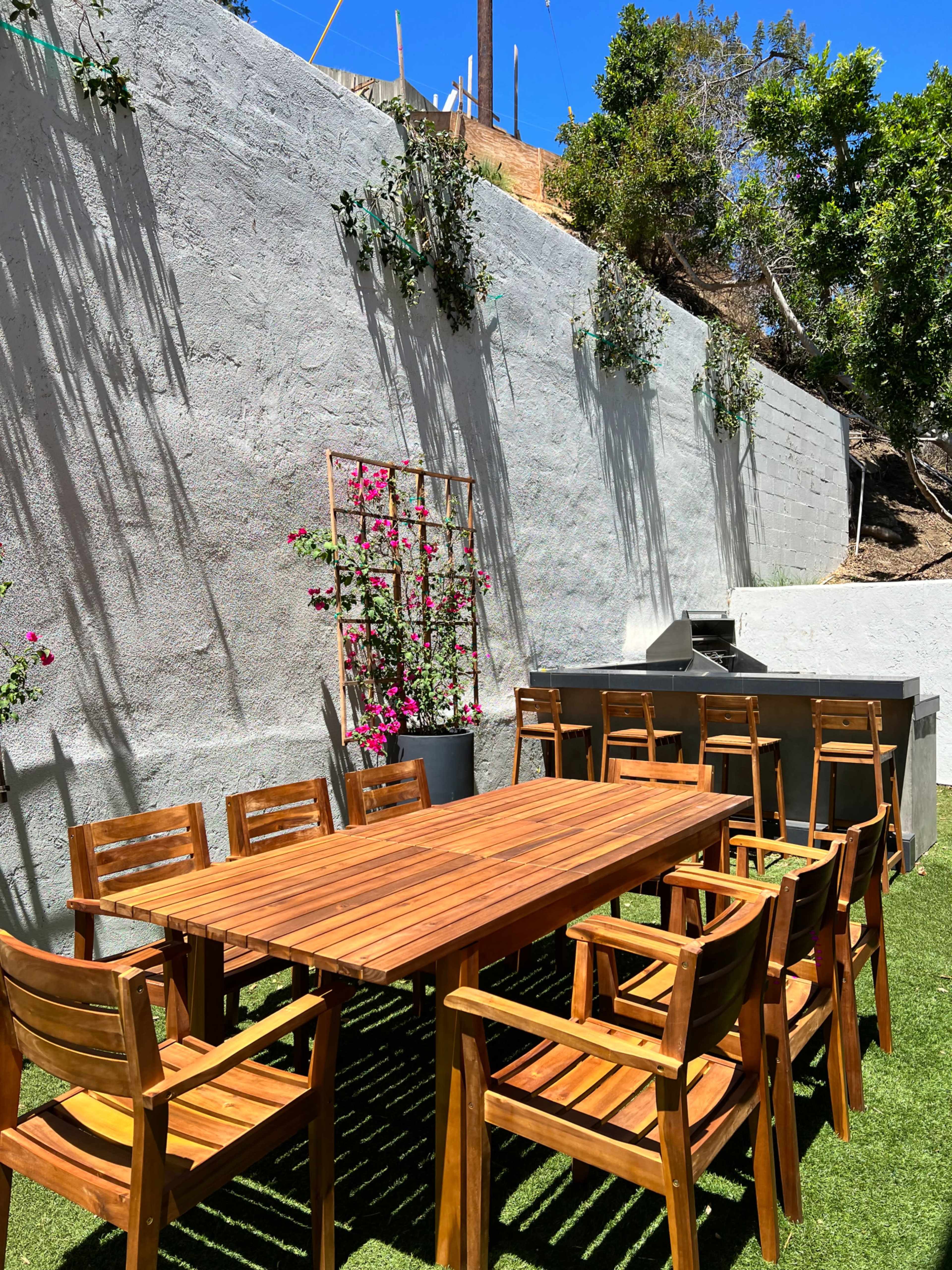 A wooden dining table surrounded by chairs is set on a grassy area next to a gray wall adorned with vertical plants.