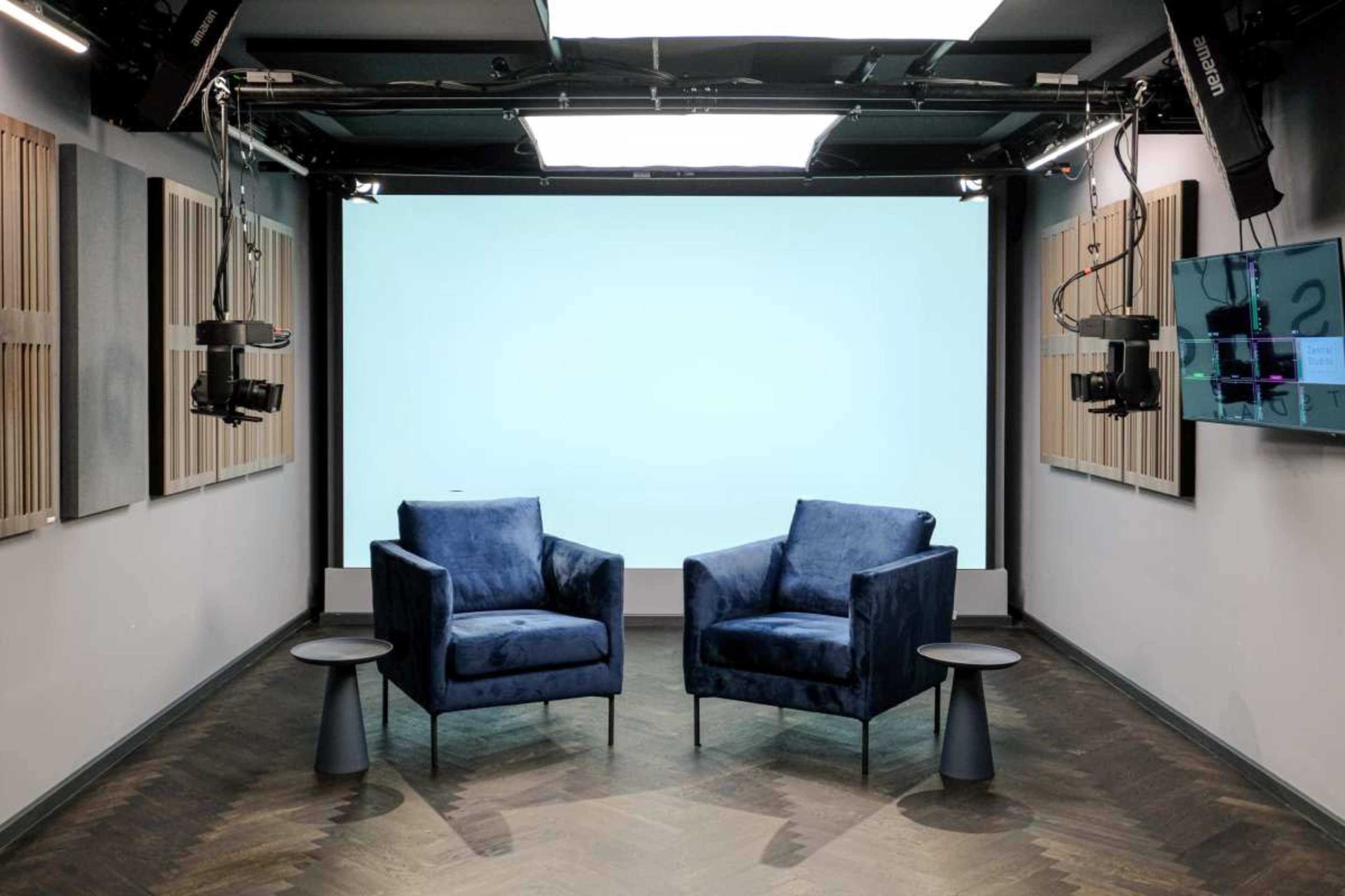 The image shows a modern studio setup featuring two blue armchairs with small side tables, against a plain light blue backdrop.
