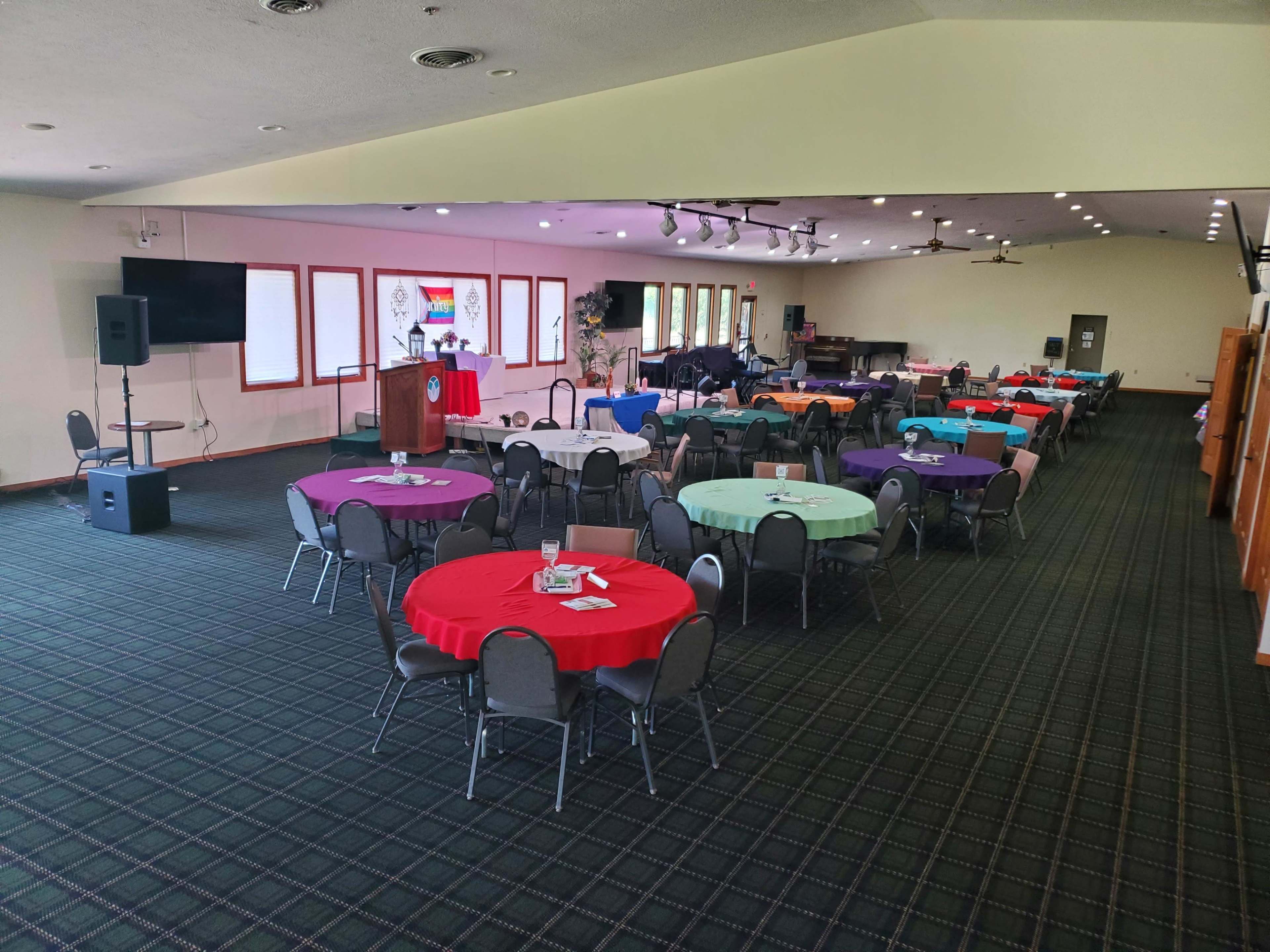 A spacious event room features multiple tables covered in colorful tablecloths arranged in rows, with a podium and a stage in the background.