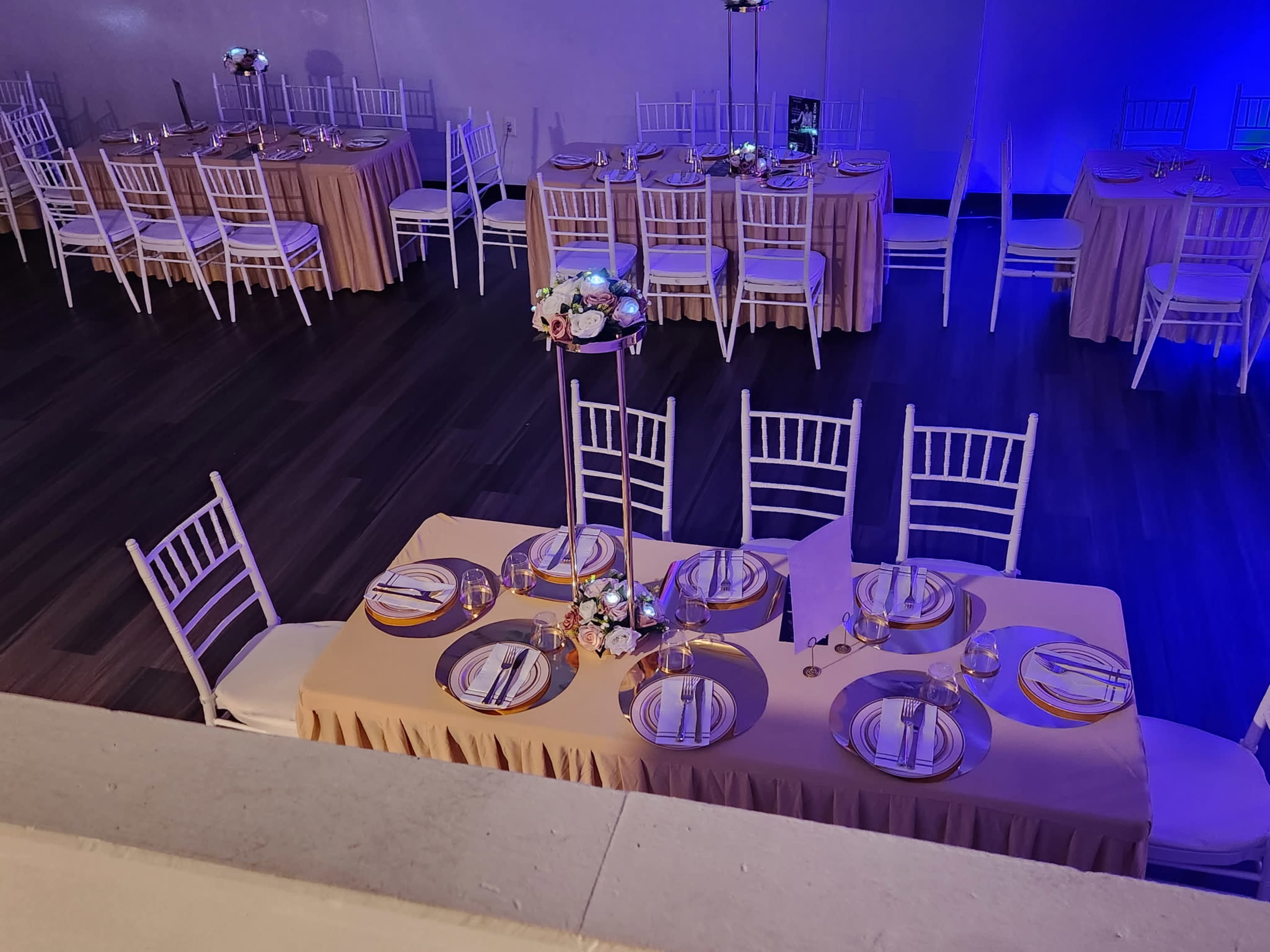 The image shows a banquet setup with tables dressed in beige cloths, white chairs, and centerpieces adorned with flowers, all arranged in a spacious, brightly lit venue.
