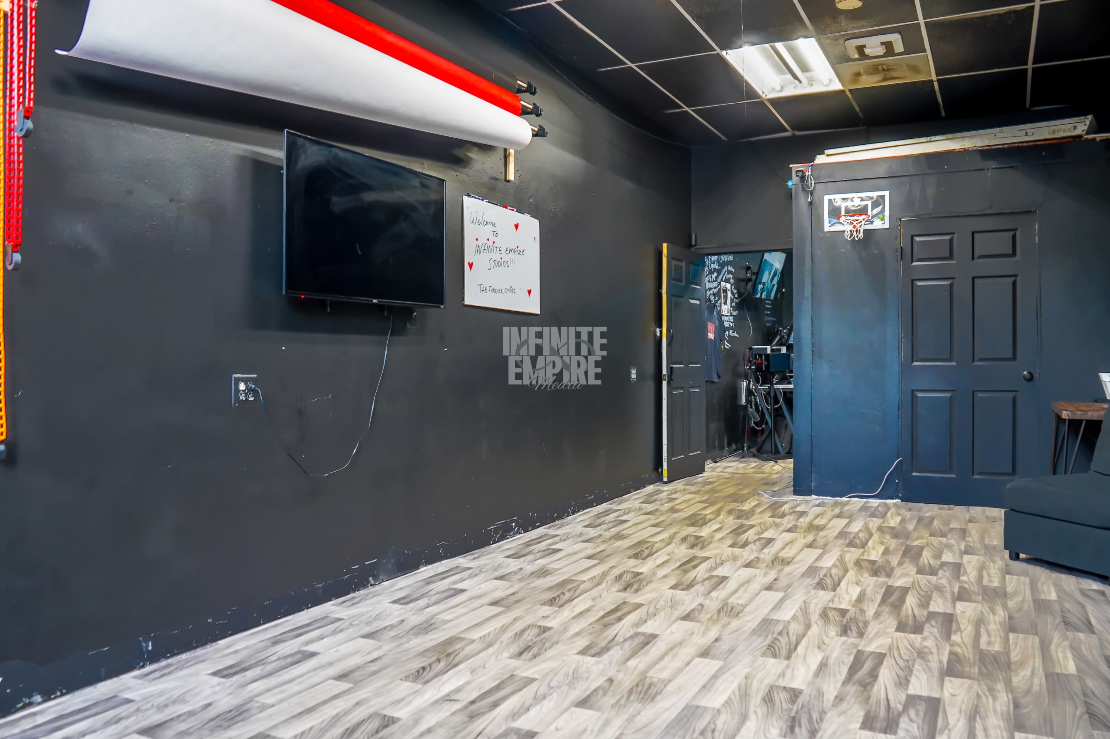 The image shows a black-walled room with a television mounted on the wall, a whiteboard with writing, and a doorway leading to another area, featuring a basketball hoop in the corner.