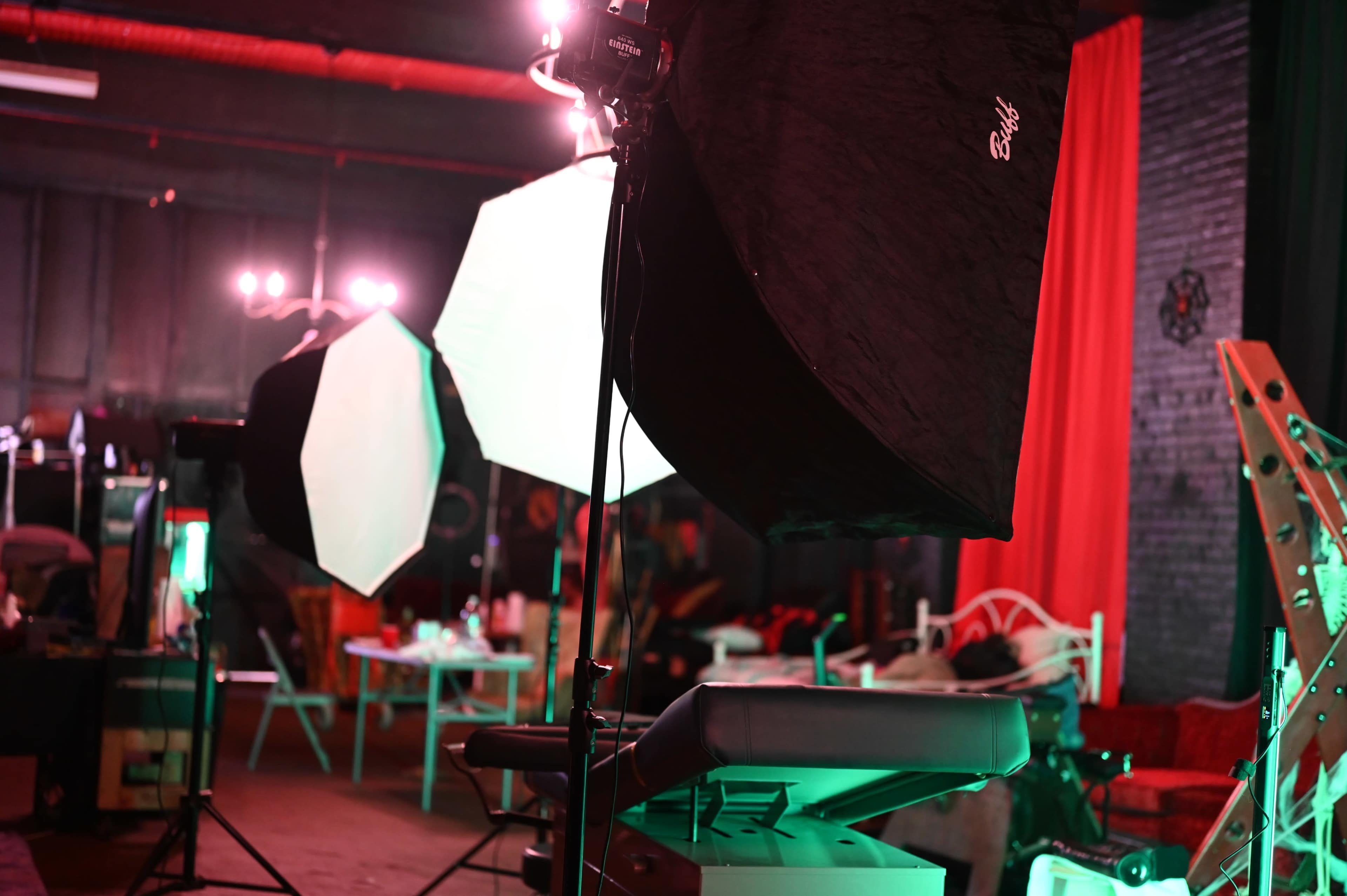 The image shows a photography studio with lighting equipment, including softboxes, set against a backdrop of red curtains and scattered furniture.