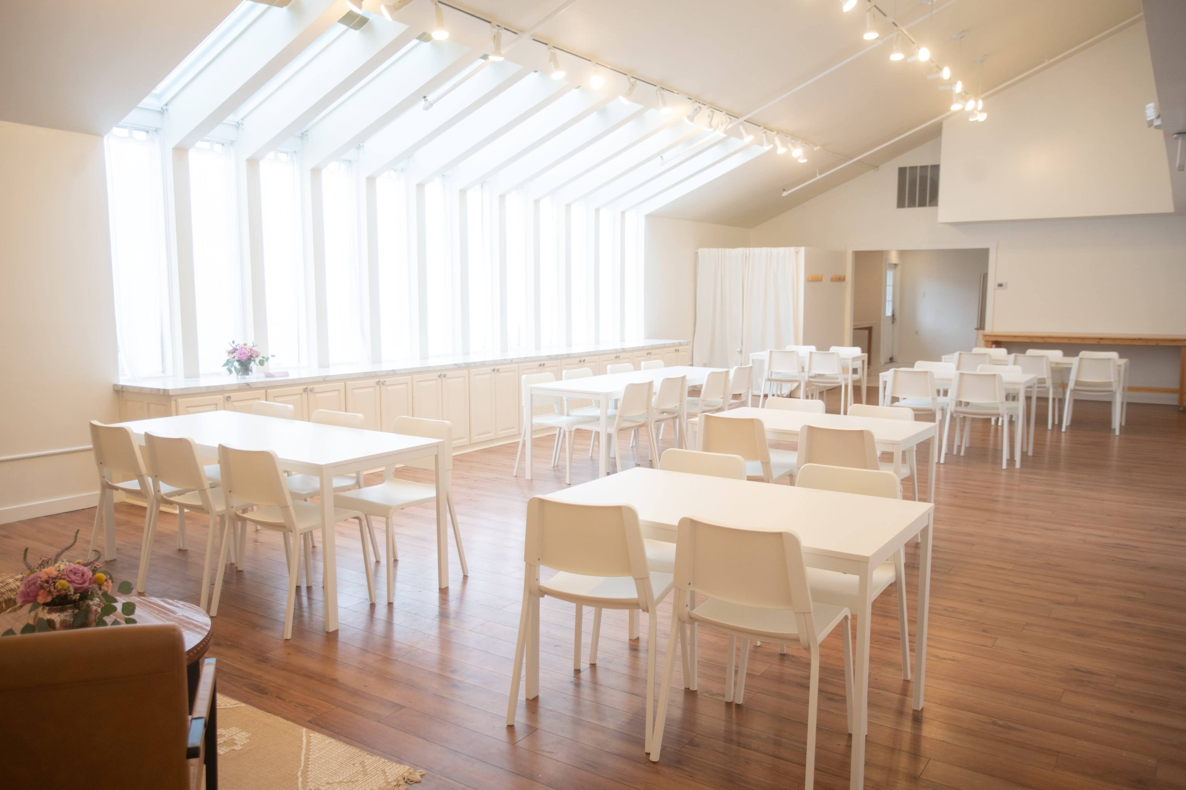 The interior space features several rows of white tables and chairs, with large windows that allow natural light to fill the room.