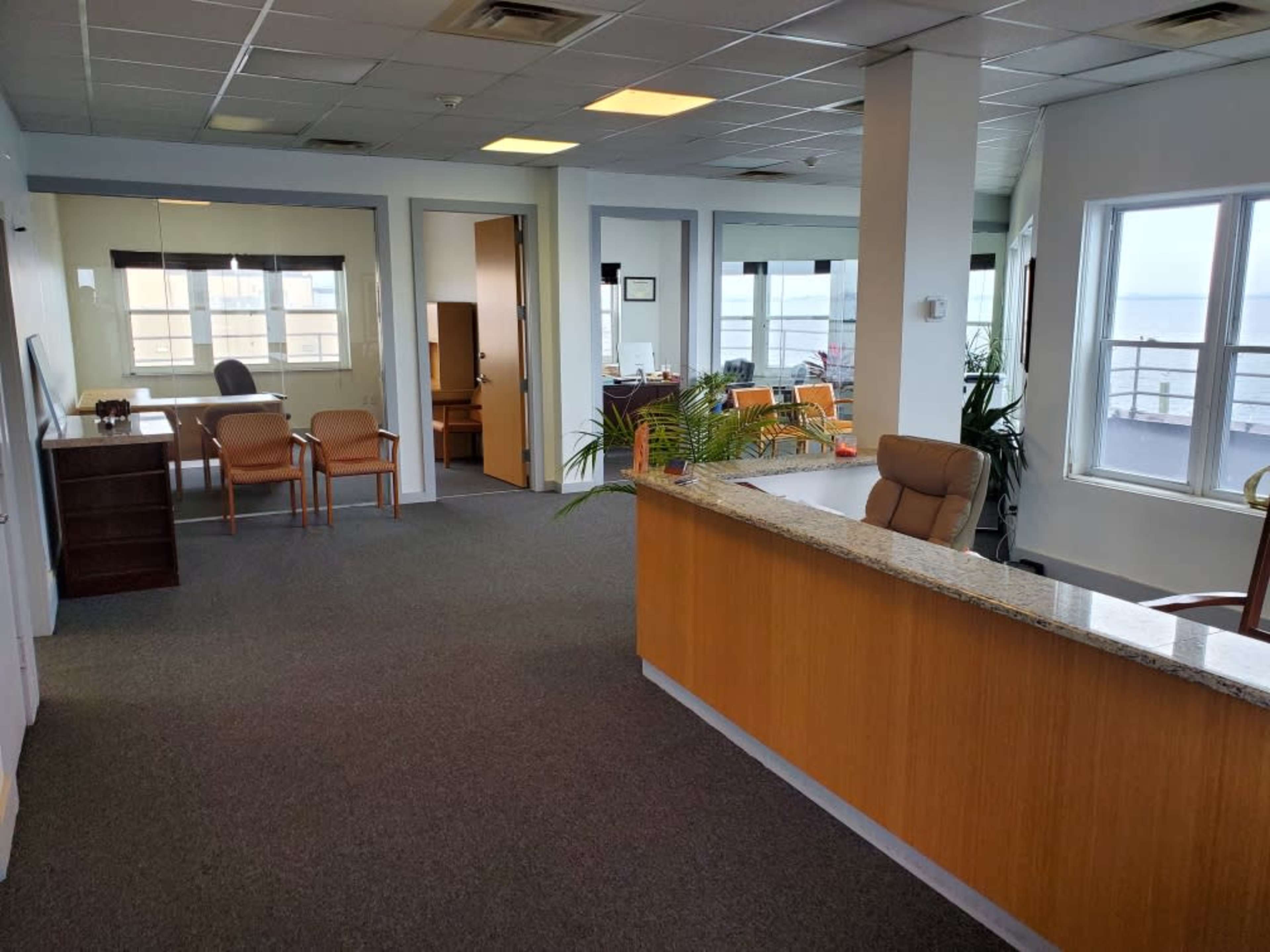 A modern office space with a reception area featuring a wooden front desk, a seating area with chairs, and large windows letting in natural light.