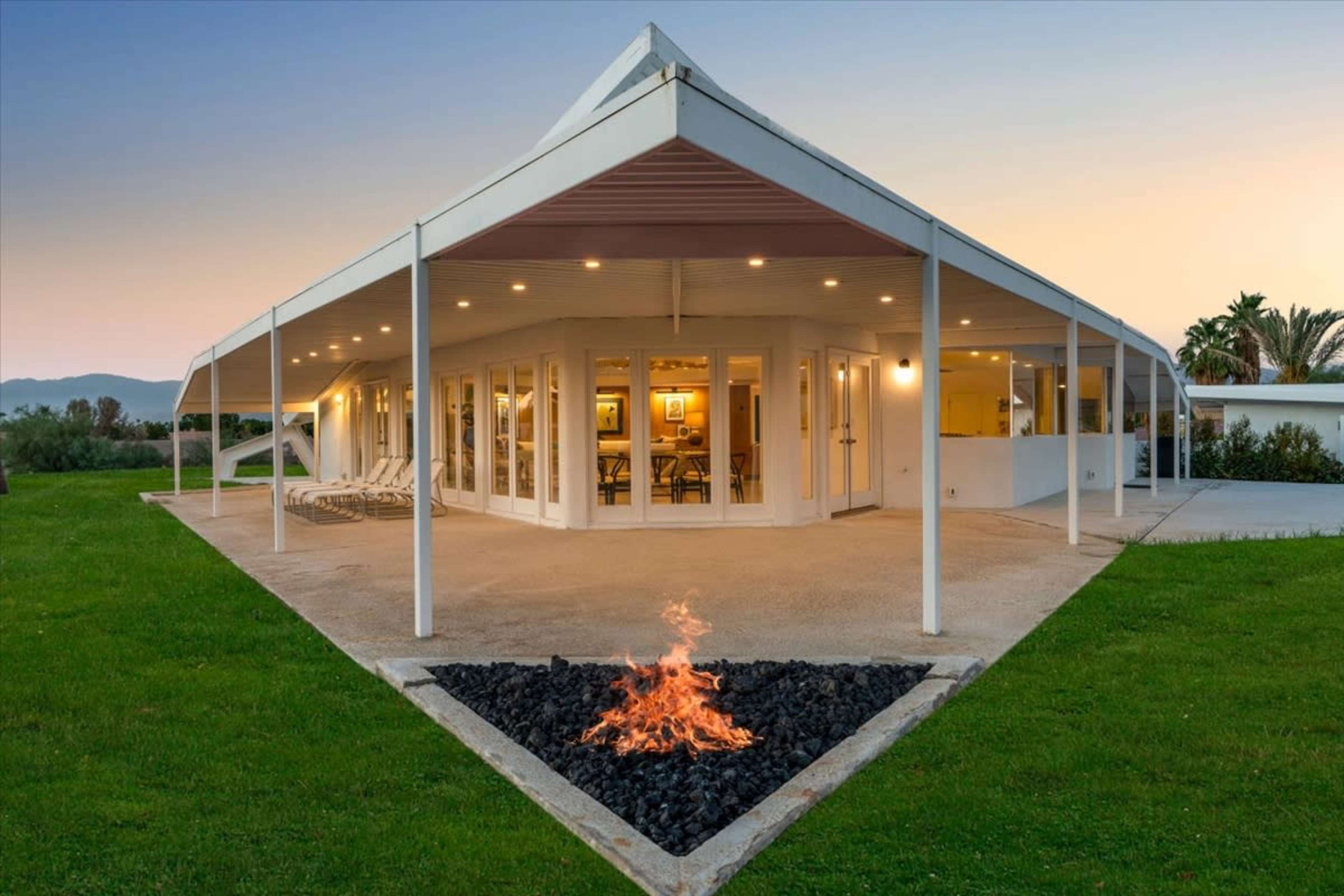 A modern house with a large covered patio and a fire pit surrounded by grass.
