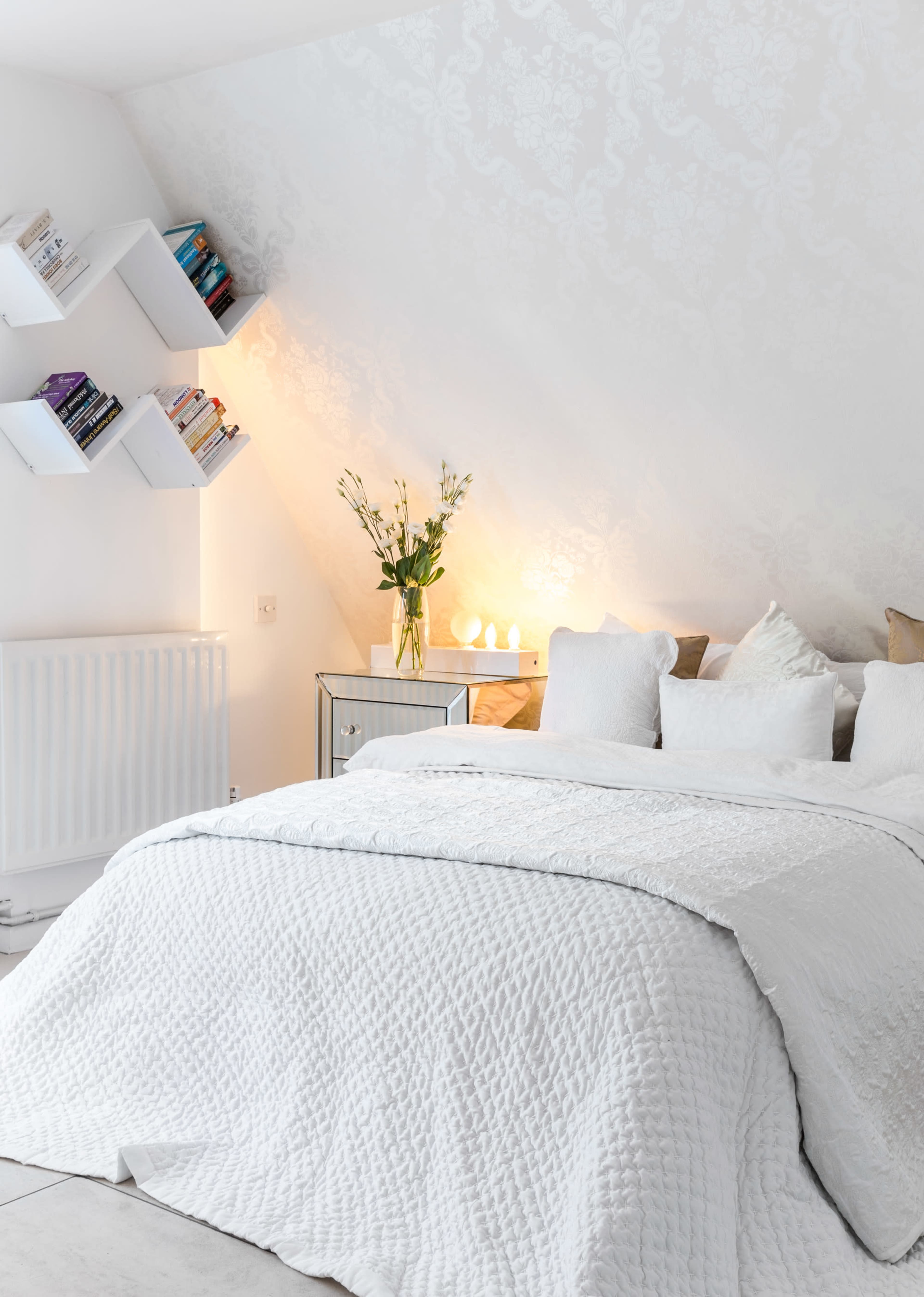 A well-decorated bedroom features a white quilted bedspread, decorative pillows, and wall-mounted bookshelves in a bright, softly lit space.