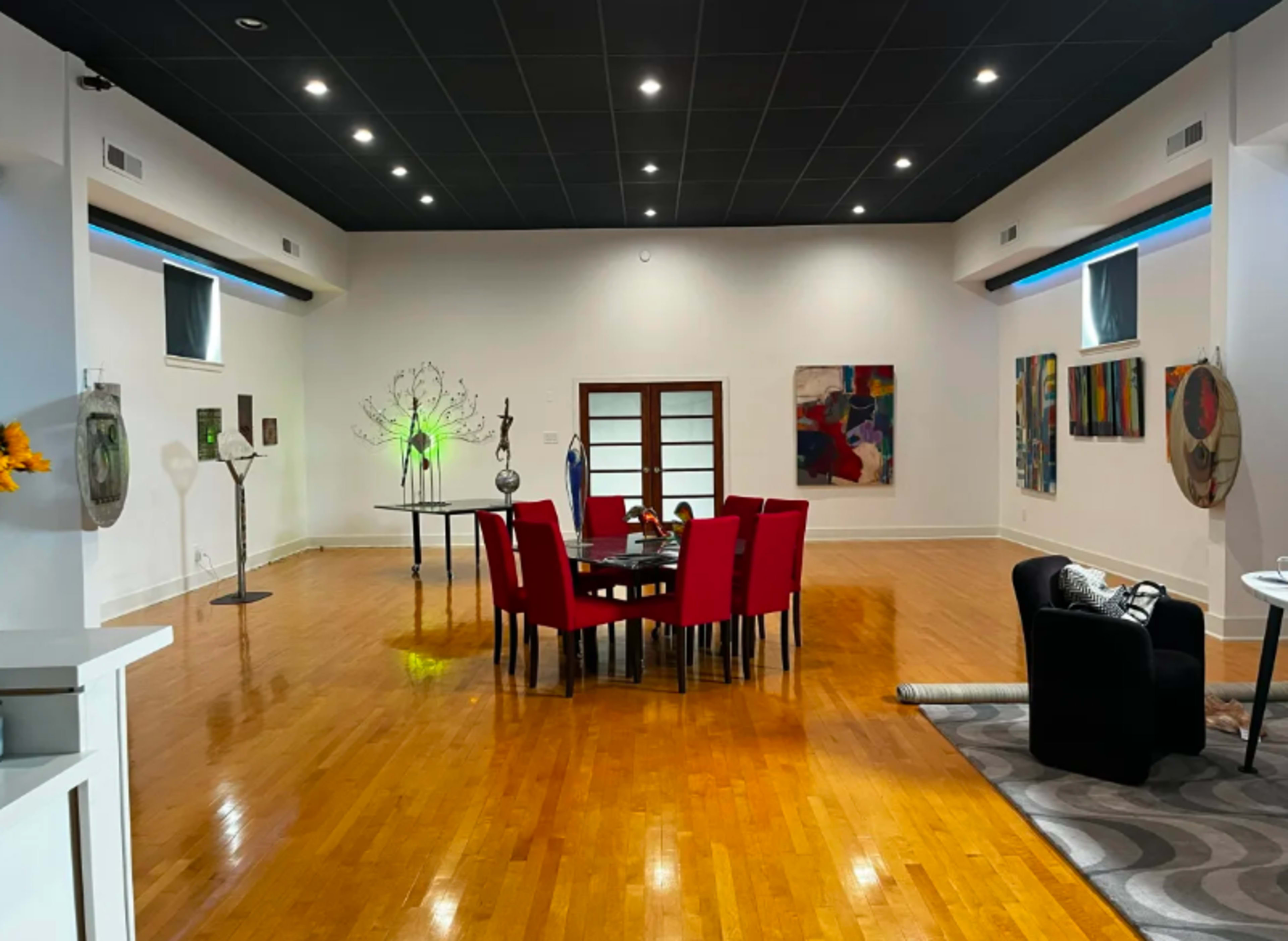 A spacious dining area features a large round table surrounded by red chairs, with various pieces of modern art on the walls and decorative objects throughout the room.