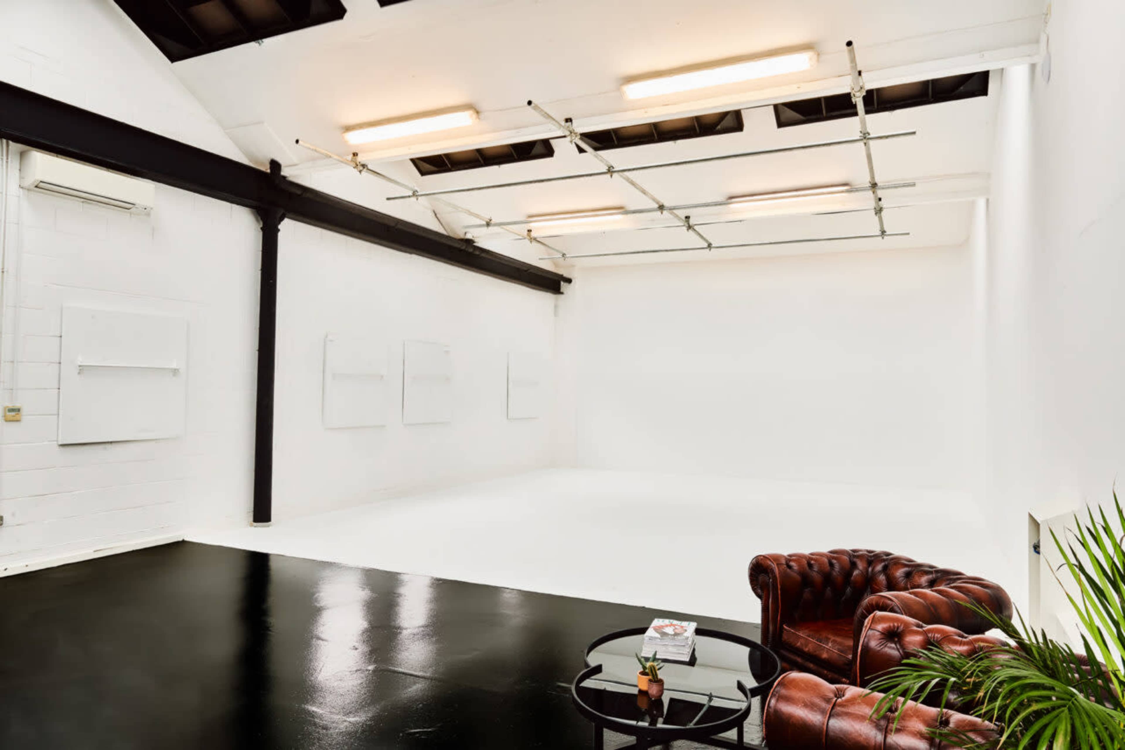 A minimalist studio space with white walls, a black floor, a brown leather chesterfield sofa, and a glass coffee table.