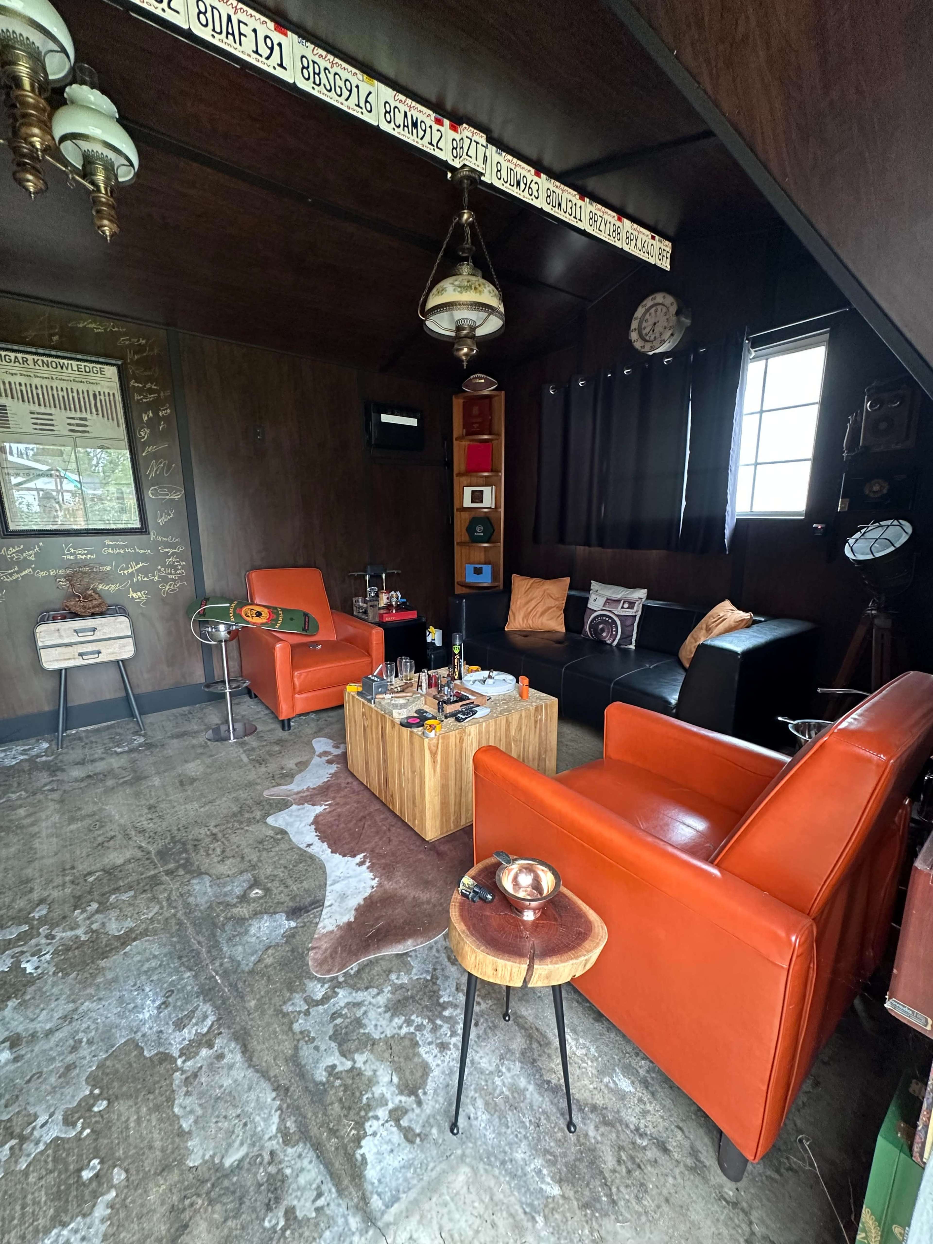 The interior of a cozy room features orange armchairs, a black sofa, and a wooden coffee table on a textured floor, with decorative items and wall art visible.