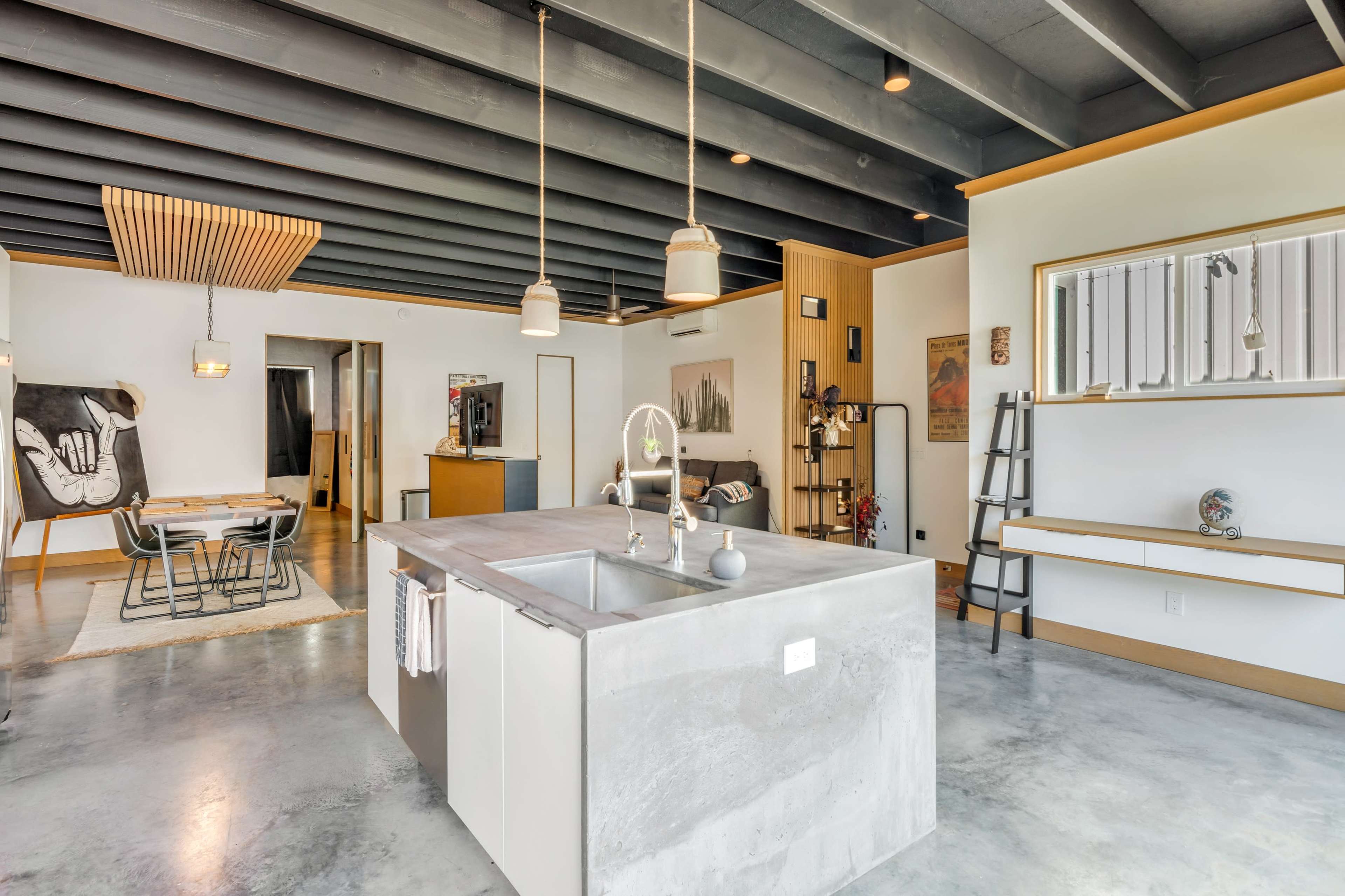A modern kitchen with a concrete island, pendant lighting, and an open layout leading to a dining area and living space.