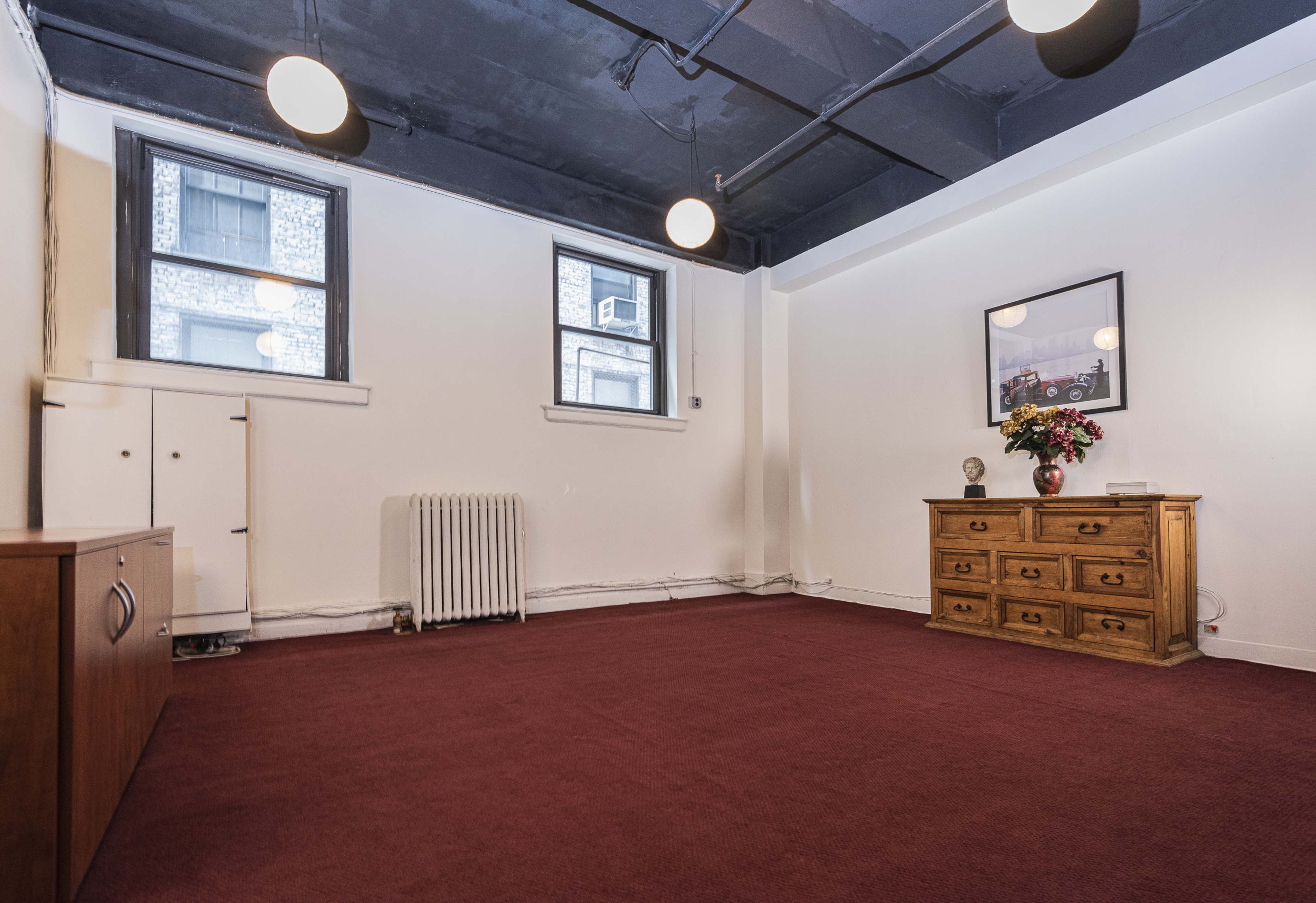 A spacious room with carpeted flooring, two windows, and a wooden cabinet with drawers and a vase of flowers on a side table.