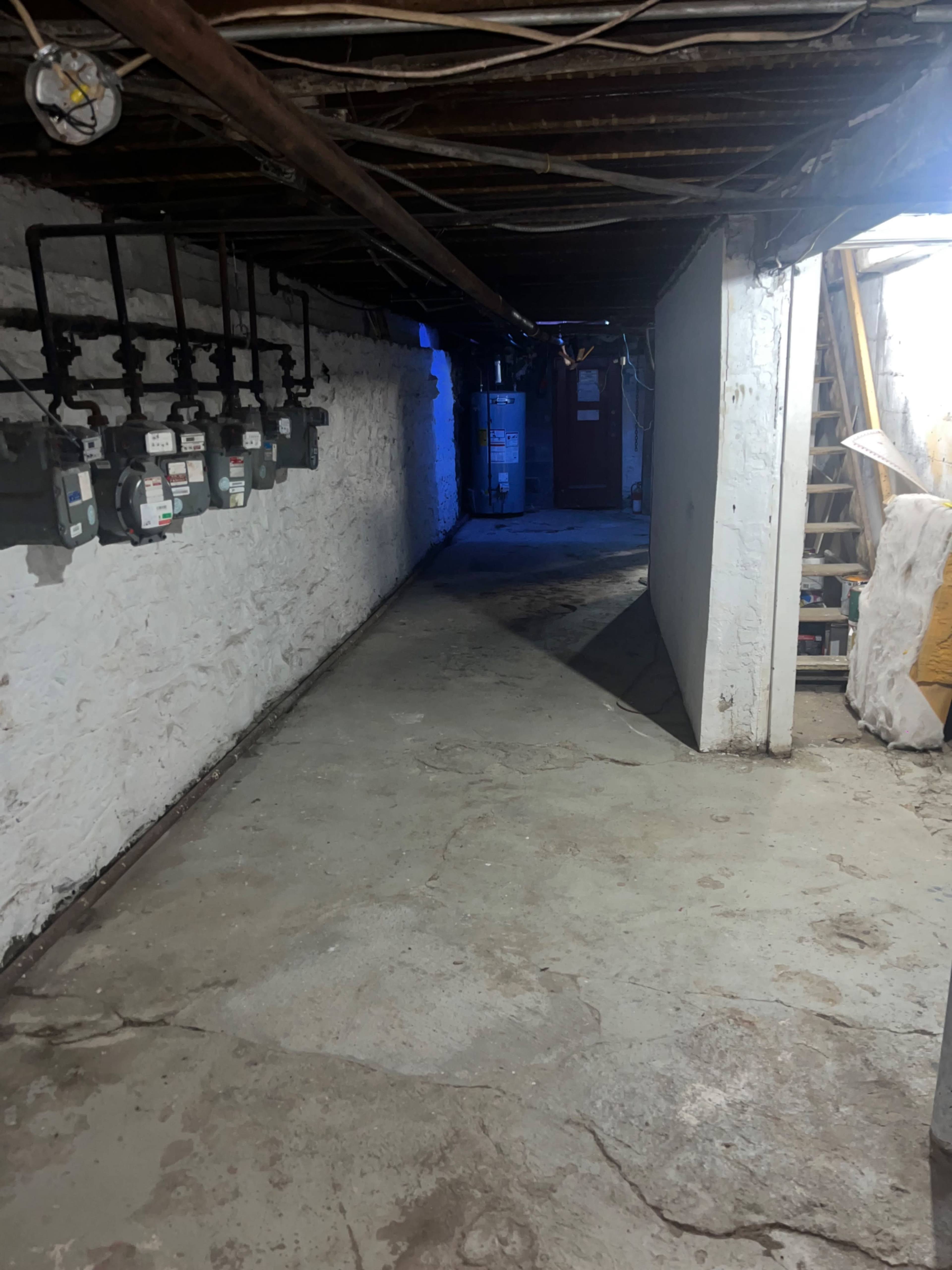The image shows a dimly lit basement hallway with exposed beams, a concrete floor, and utility meters mounted on the left wall.