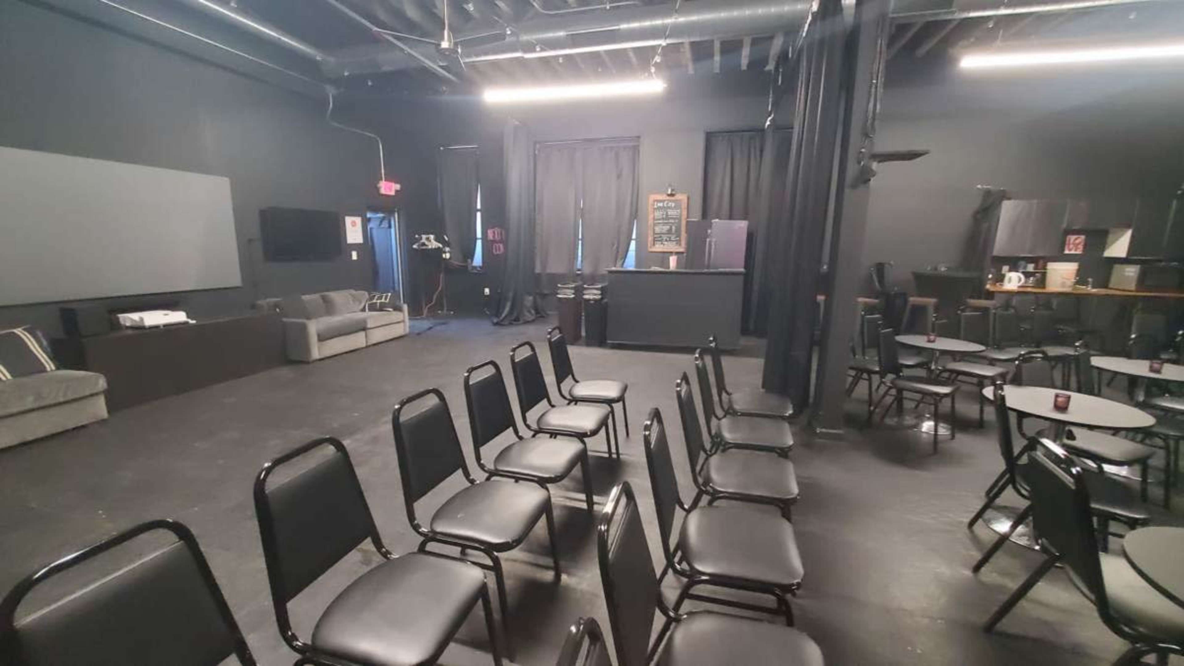 The image shows a black-walled room set up for an event, featuring rows of metal chairs facing a bar area with a chalkboard menu.