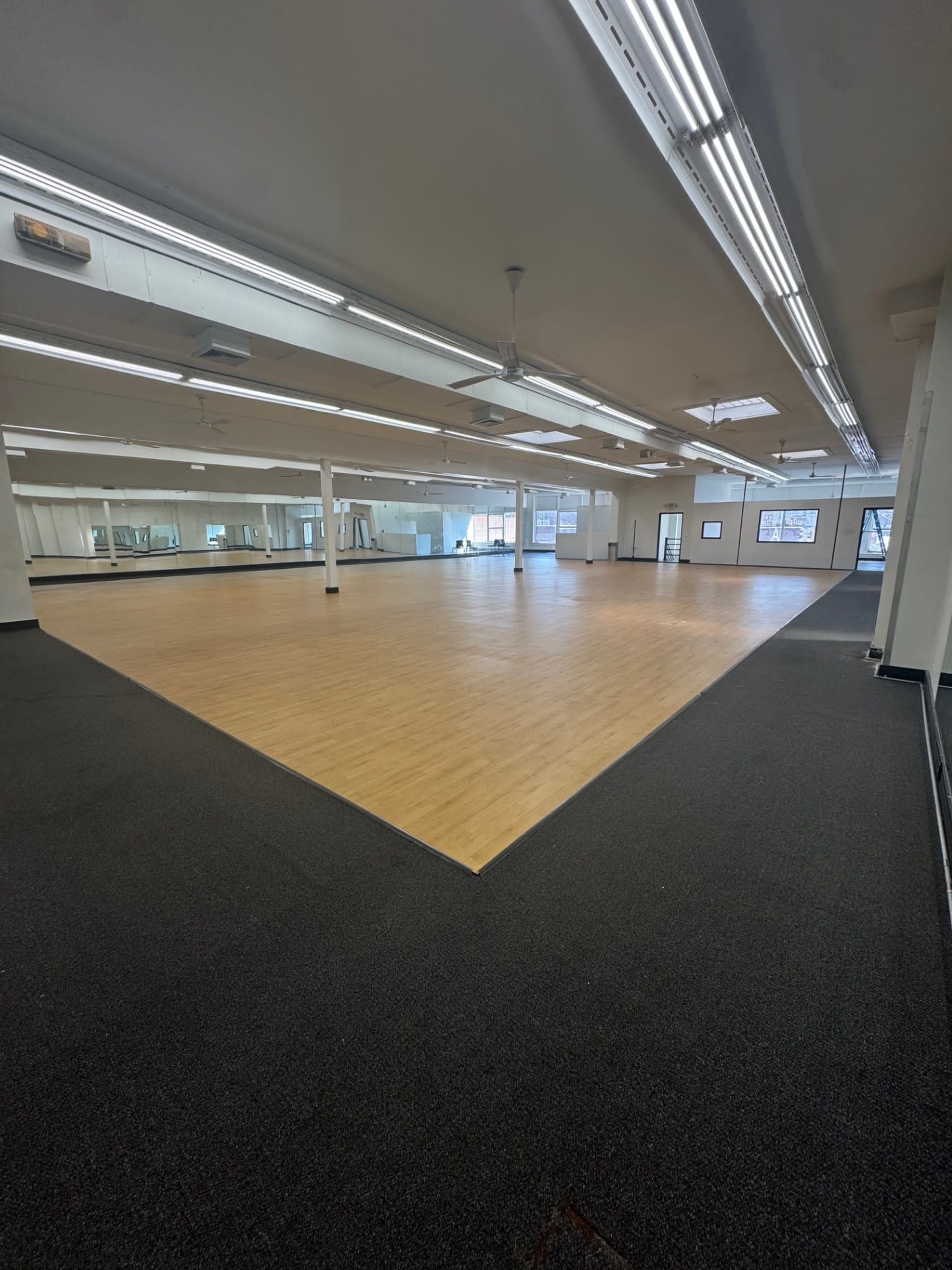 The image shows a spacious, empty room with wooden flooring and large mirrors along the walls.