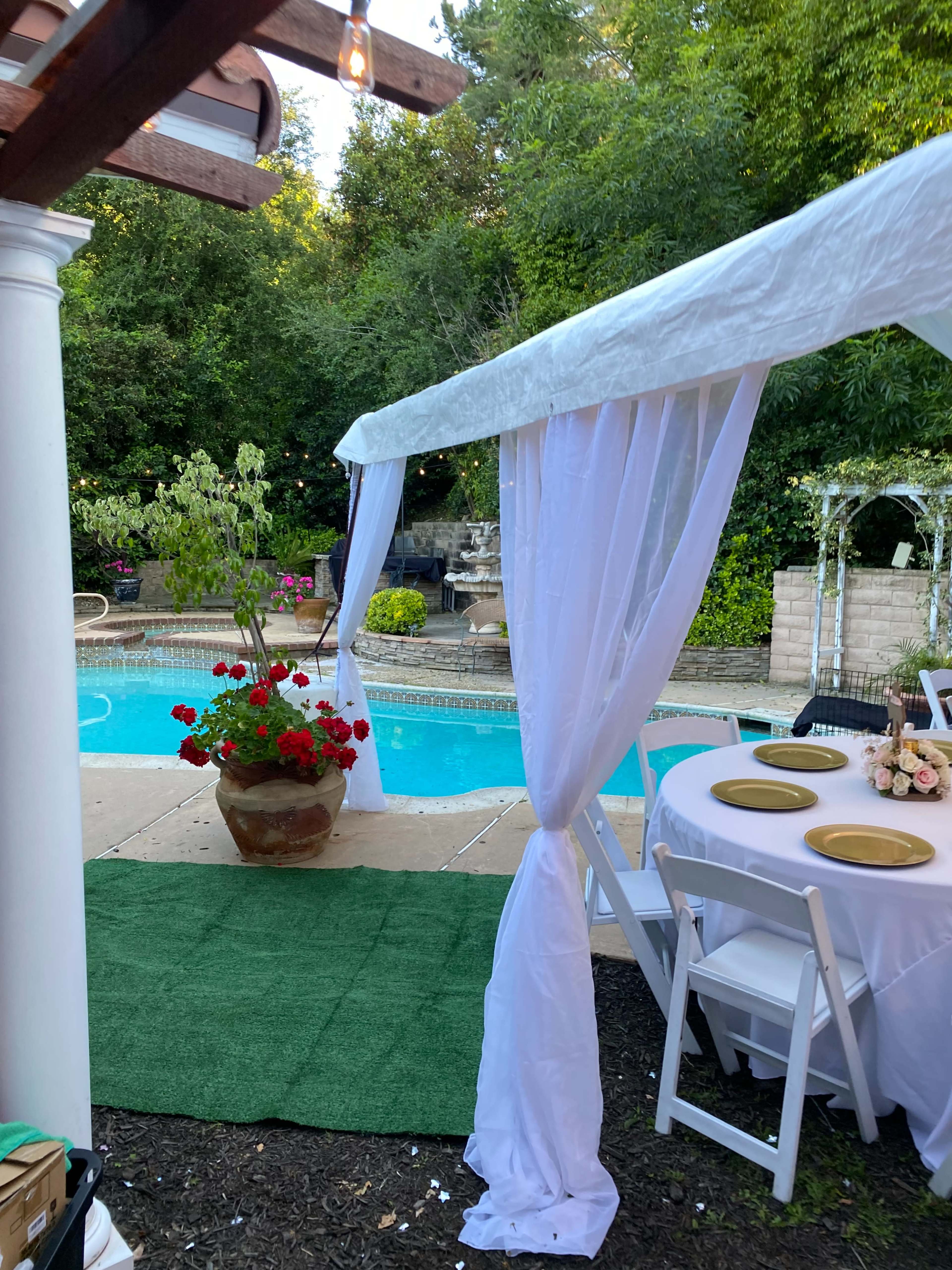 A decorated outdoor space features a pool surrounded by greenery, with a table set for a gathering and a large flower pot in the foreground.