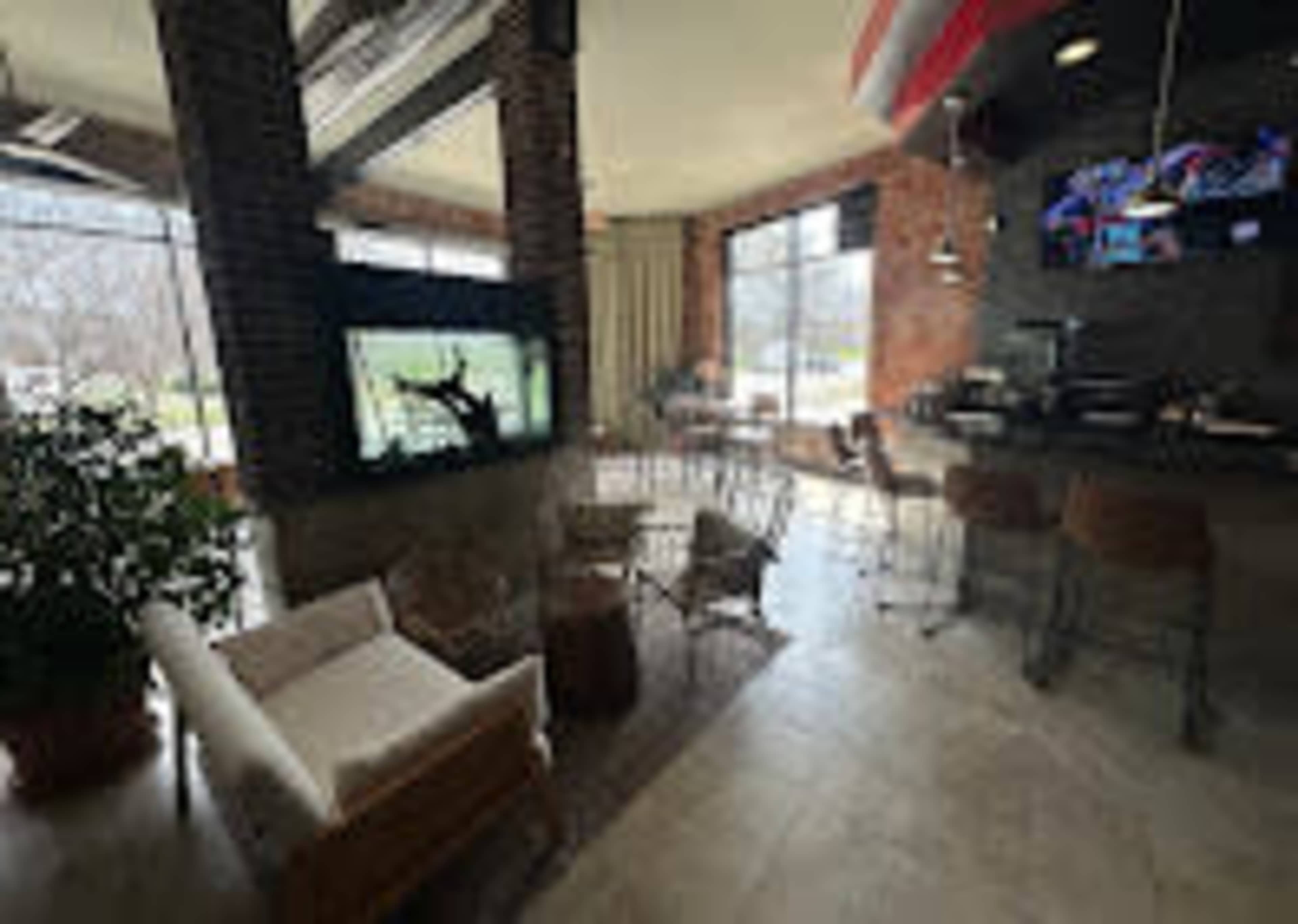 The image shows a modern café interior with a seating area, large windows allowing natural light, and a television screen mounted on a wall.