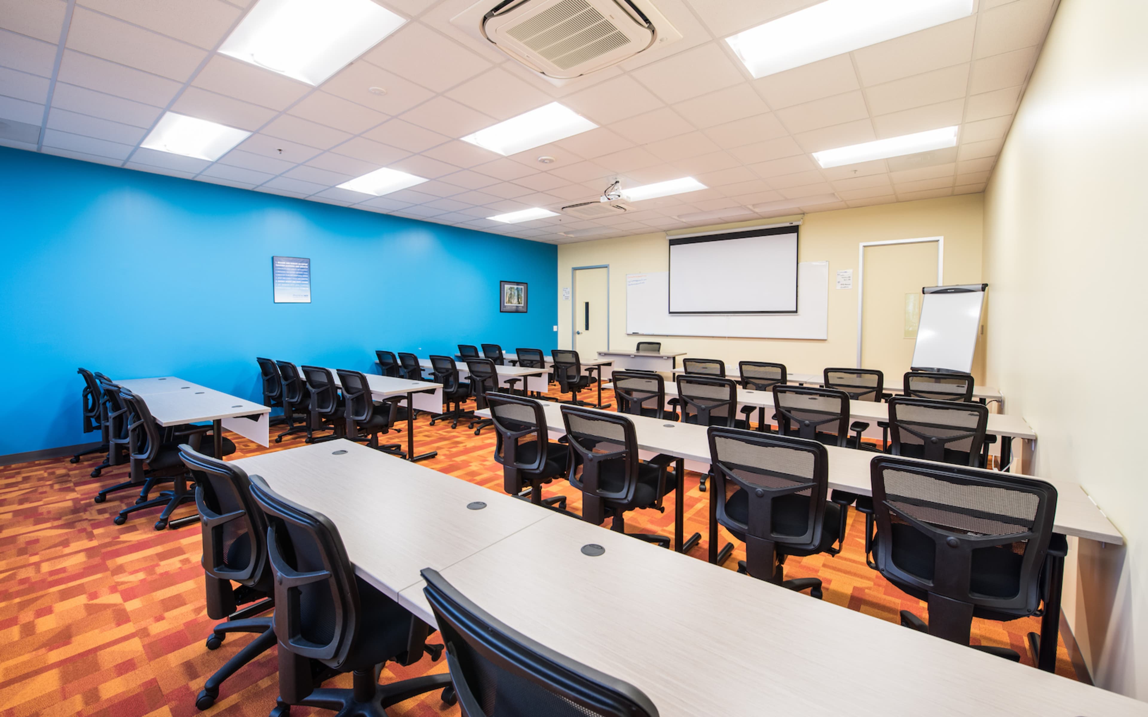 The image shows a classroom setting with several rows of tables and chairs, a projector screen at the front, and brightly colored walls.