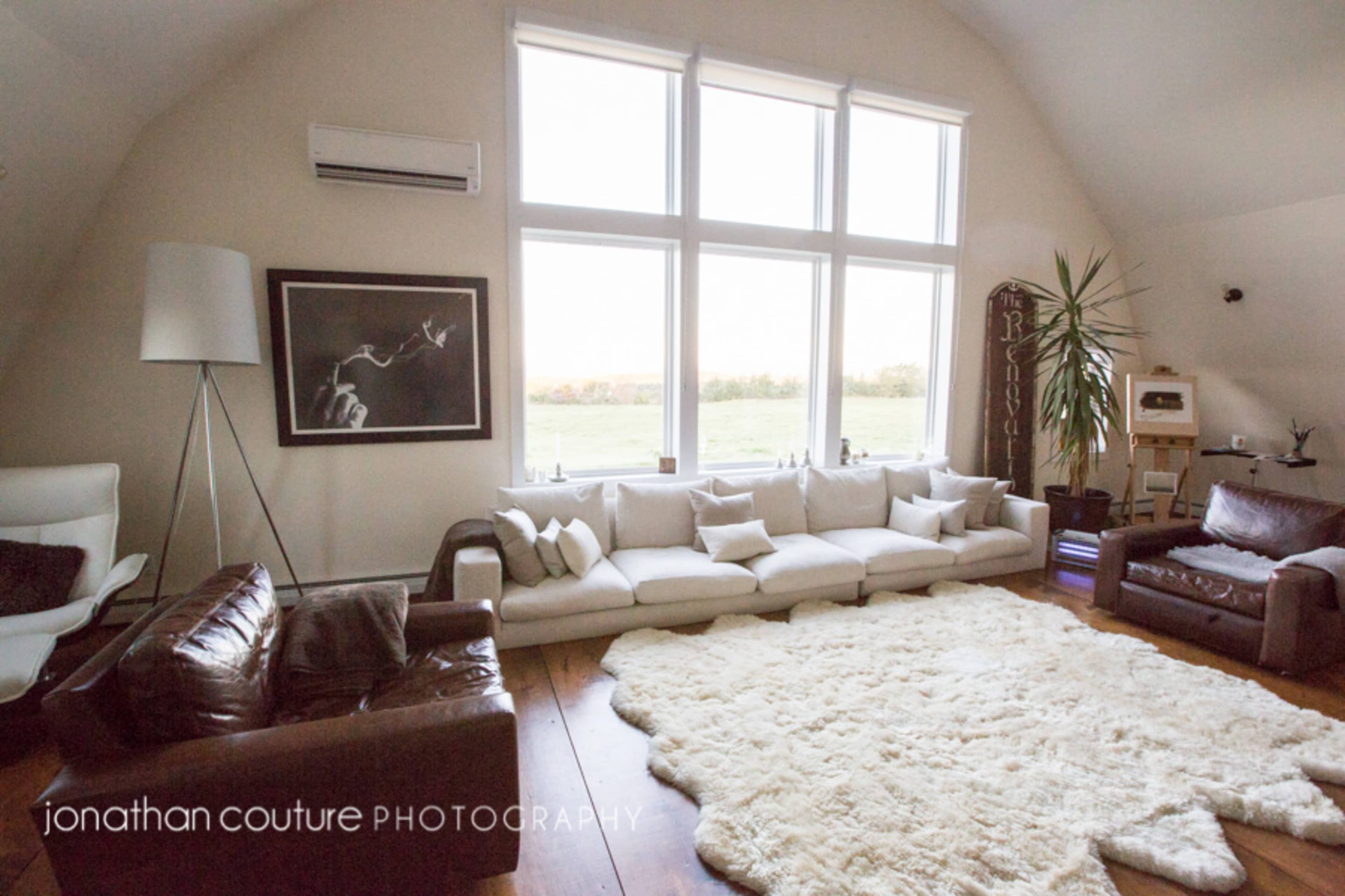 A spacious living room with large windows, a white sectional sofa, a brown leather chair, and a plush white rug on a wooden floor.