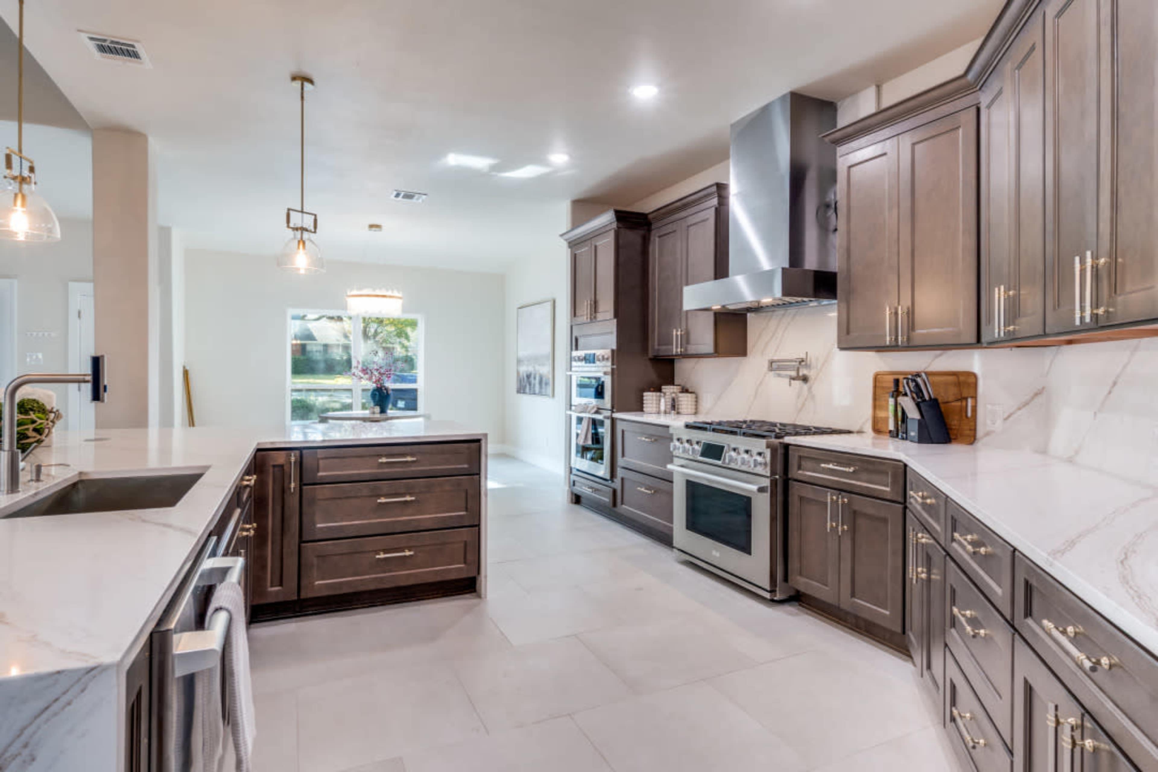 The image shows a modern kitchen featuring dark wooden cabinets, a large marble countertop, and stainless steel appliances, with natural light coming through a window.