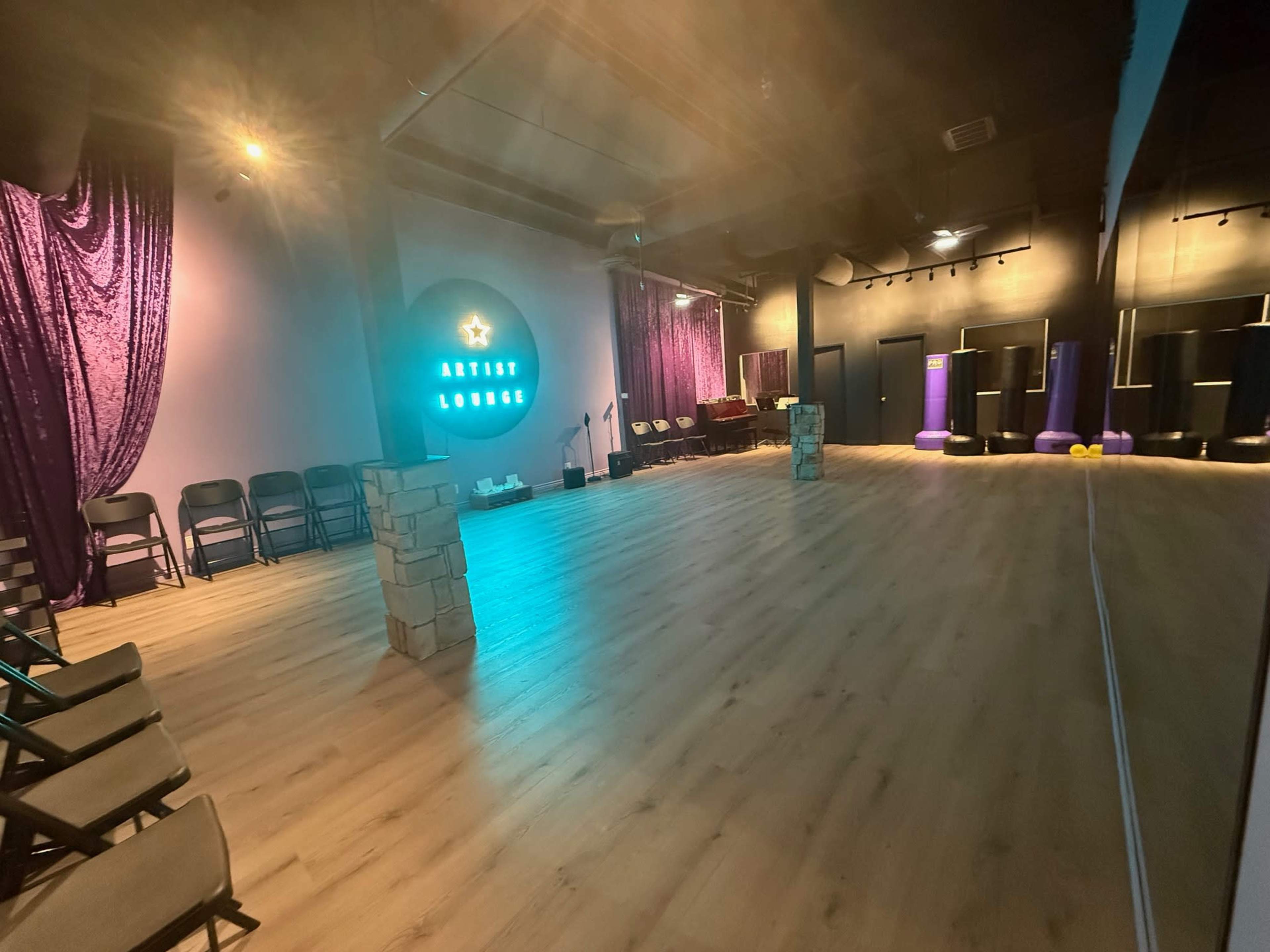 The image shows a spacious interior of an artist lounge with purple curtains, a wooden floor, and seating arranged on one side.