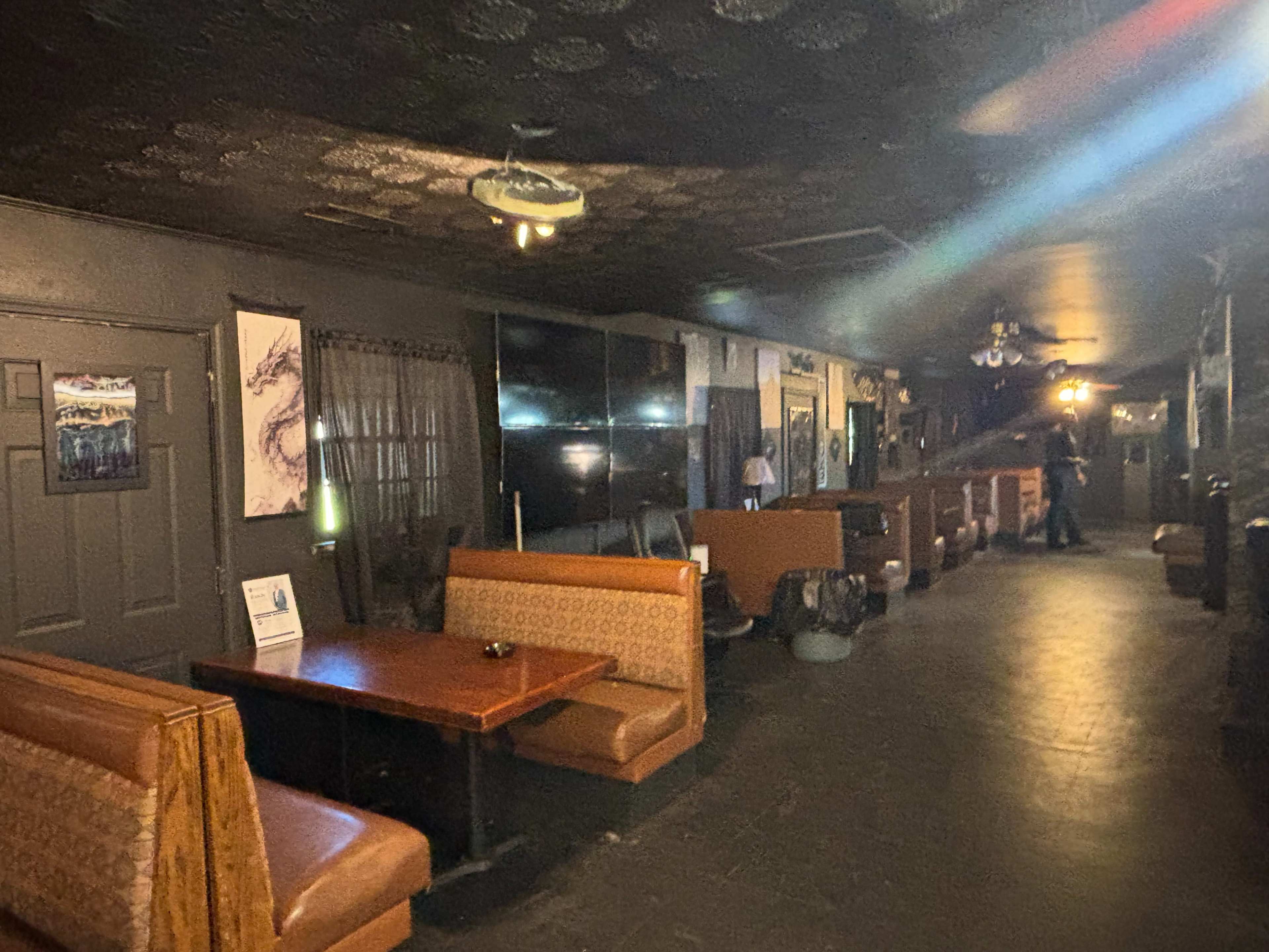 The image shows the interior of a dimly lit bar or lounge with booths and tables along one side and a large screen on the wall.