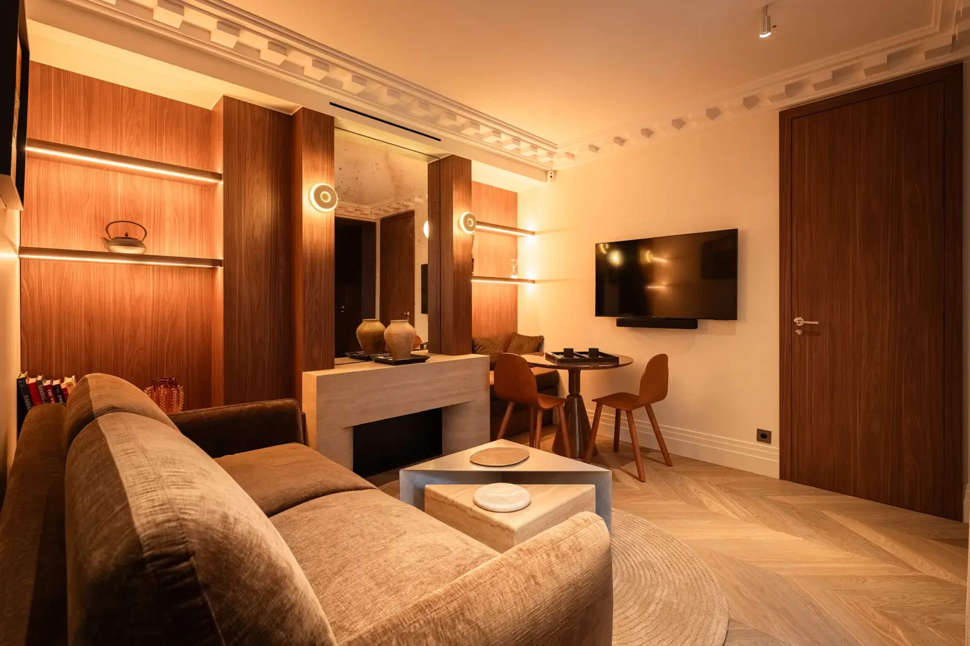 The image shows a modern living space featuring a cozy sofa, a flat-screen TV mounted on the wall, and a small dining area with wooden chairs.