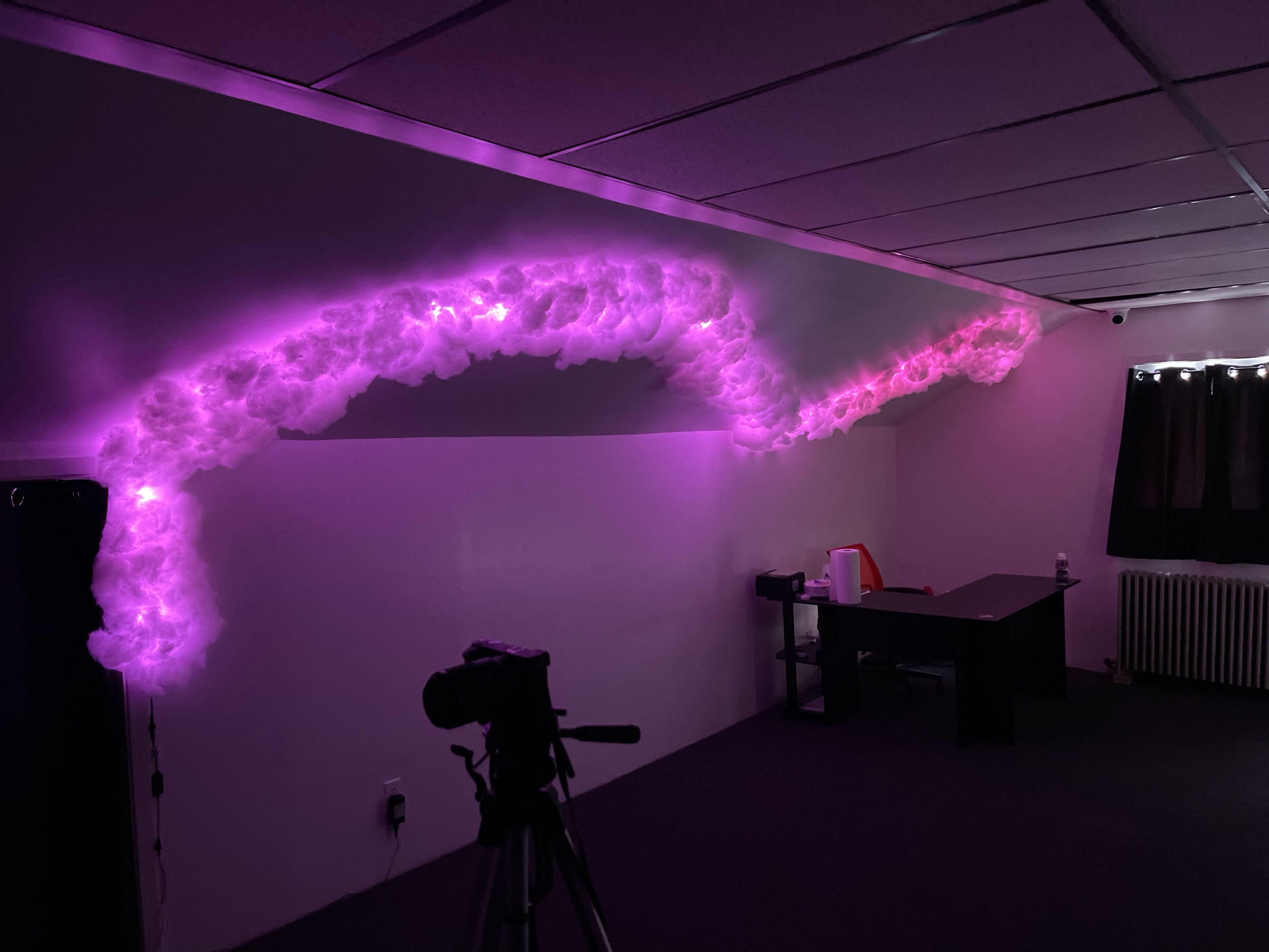 A room with a dark carpet and a desk, featuring a wall adorned with purple-lit cotton resembling clouds.