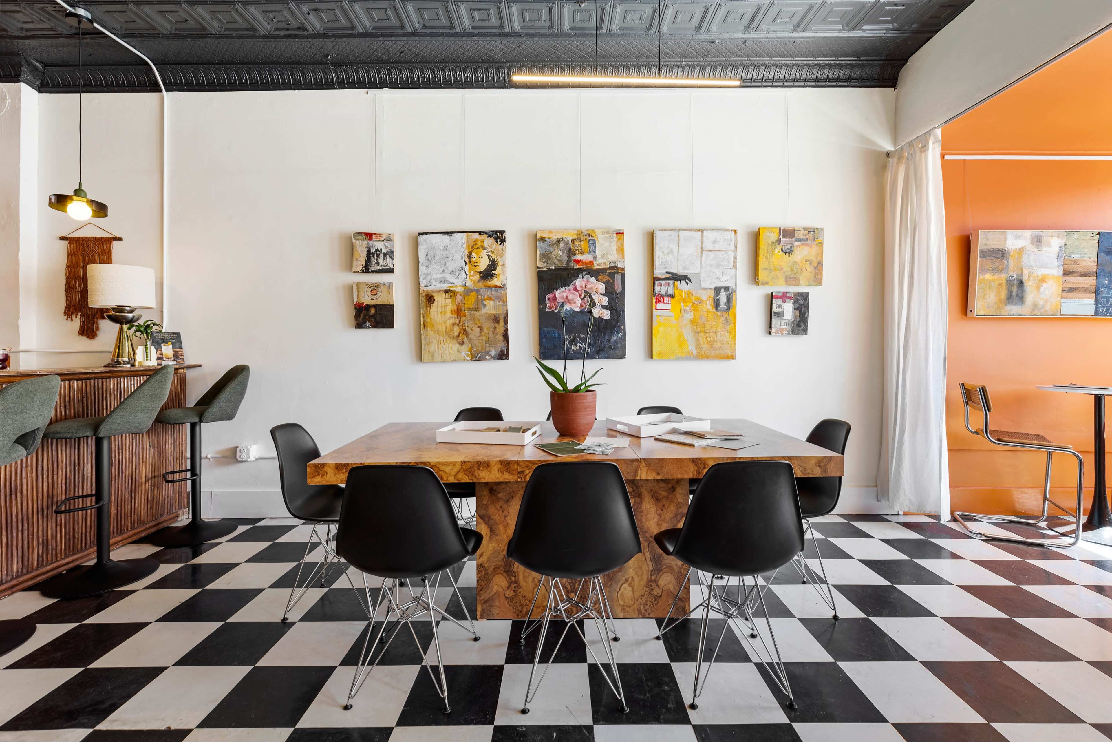 A dining area features a large wooden table surrounded by black chairs, with colorful artwork displayed on the wall and contrasting elements of a checkered floor.