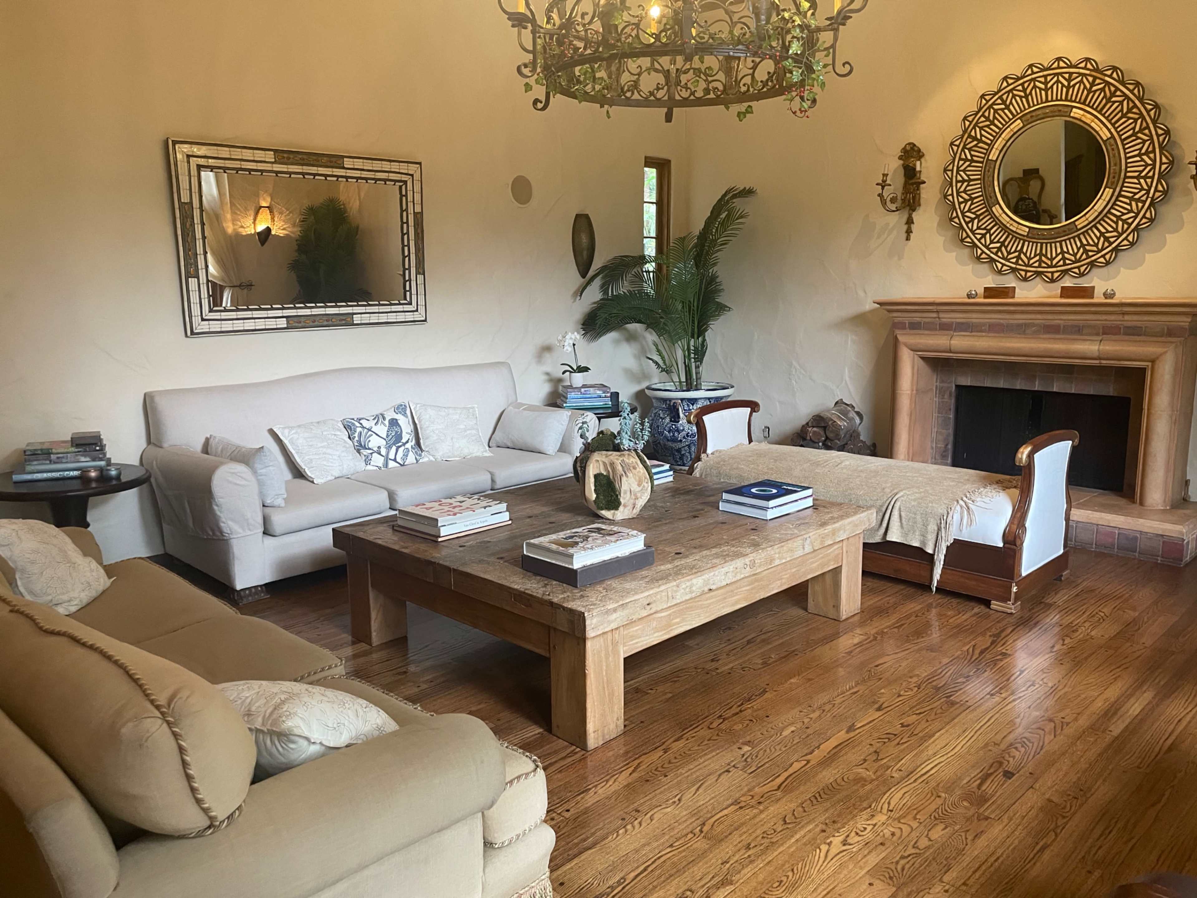 A spacious living room features a light-colored sofa, a wooden coffee table, and a fireplace, complemented by a large round mirror and potted plants.