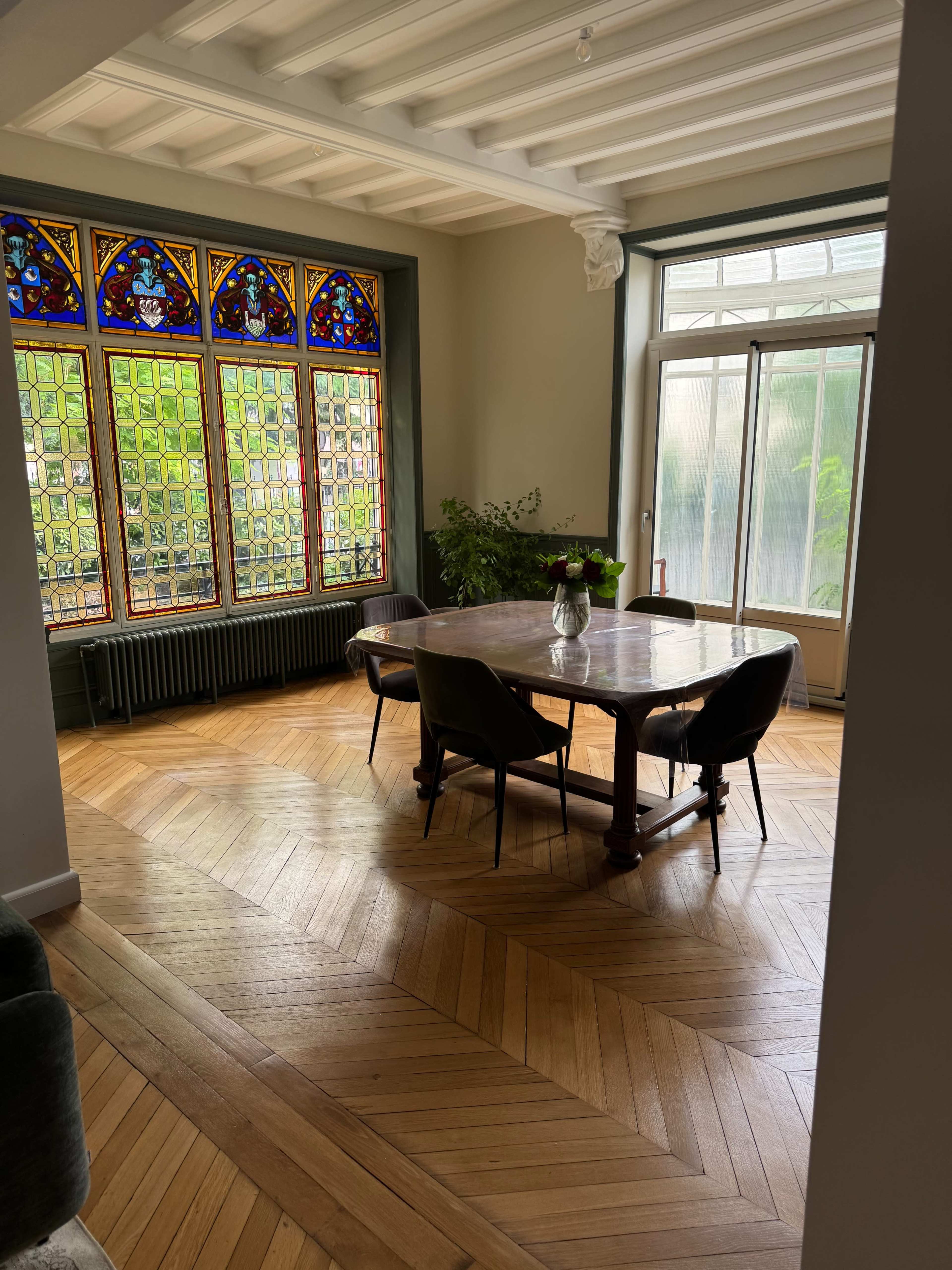 A dining area features a wooden table surrounded by black chairs, illuminated by colorful stained glass windows.