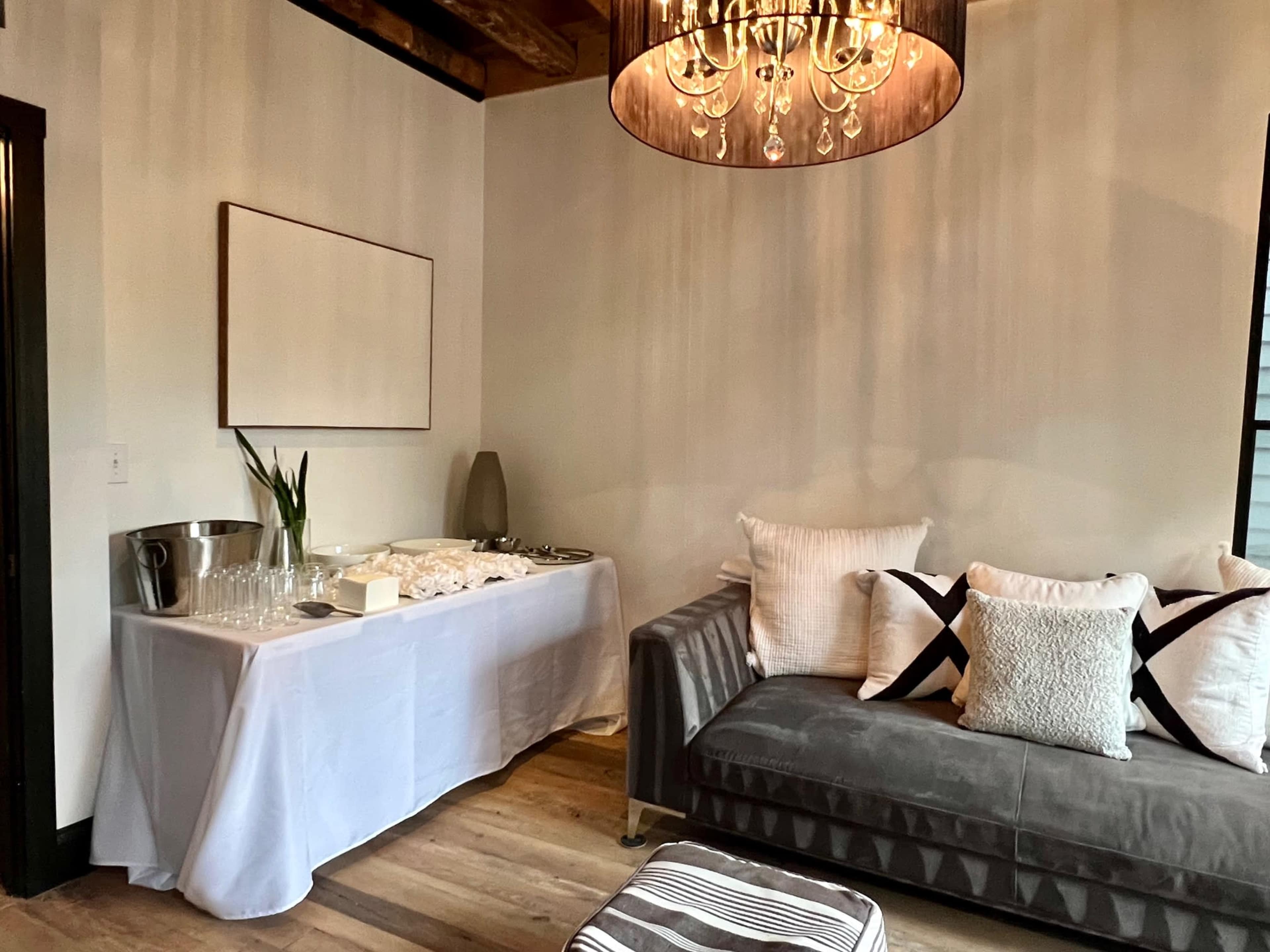 A modern living space featuring a gray sofa with decorative pillows, a white table set with glassware and a small centerpiece, and a large chandelier overhead.