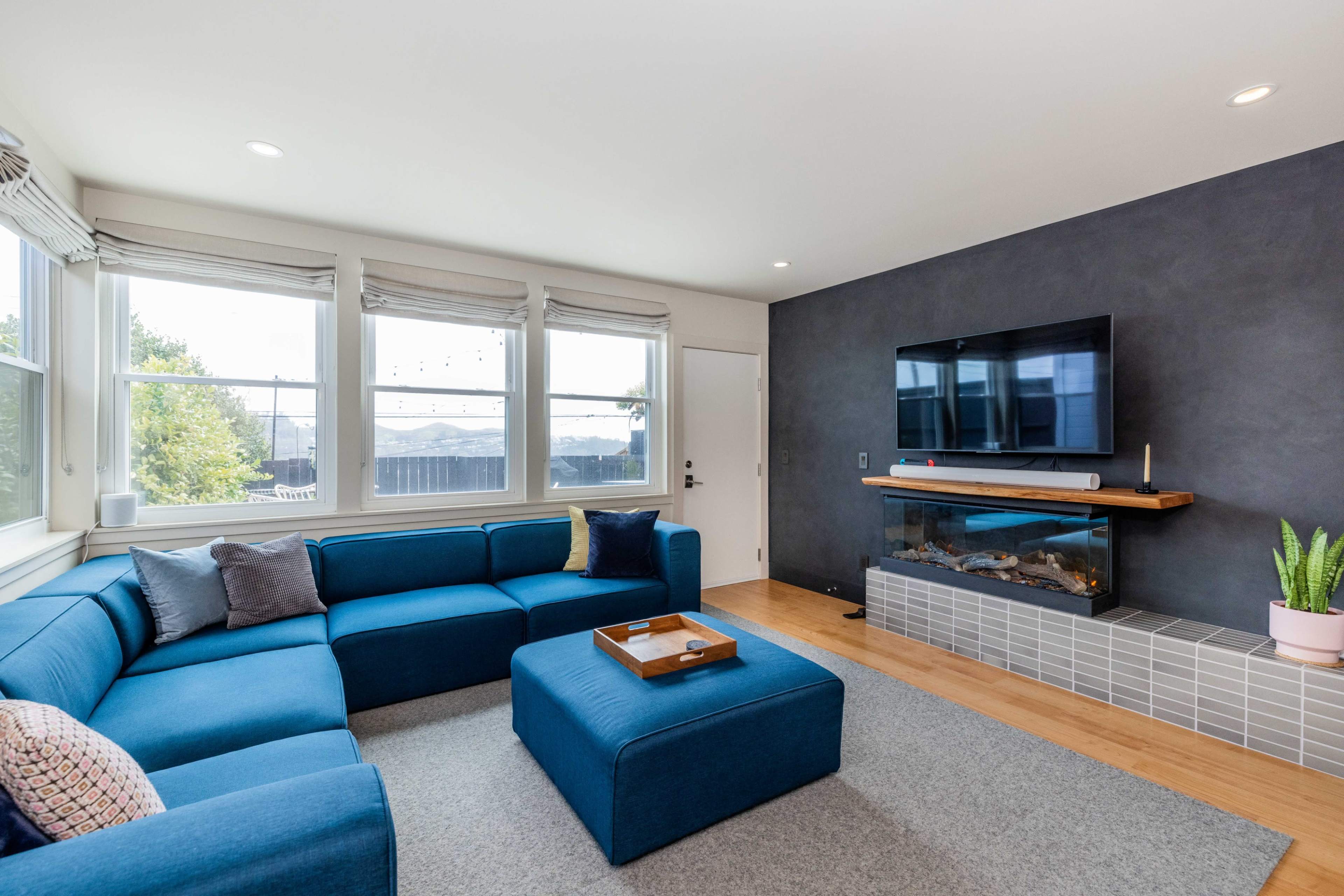 A spacious living room features a blue sectional sofa, a mounted television, and a small fireplace under a modern mantel.